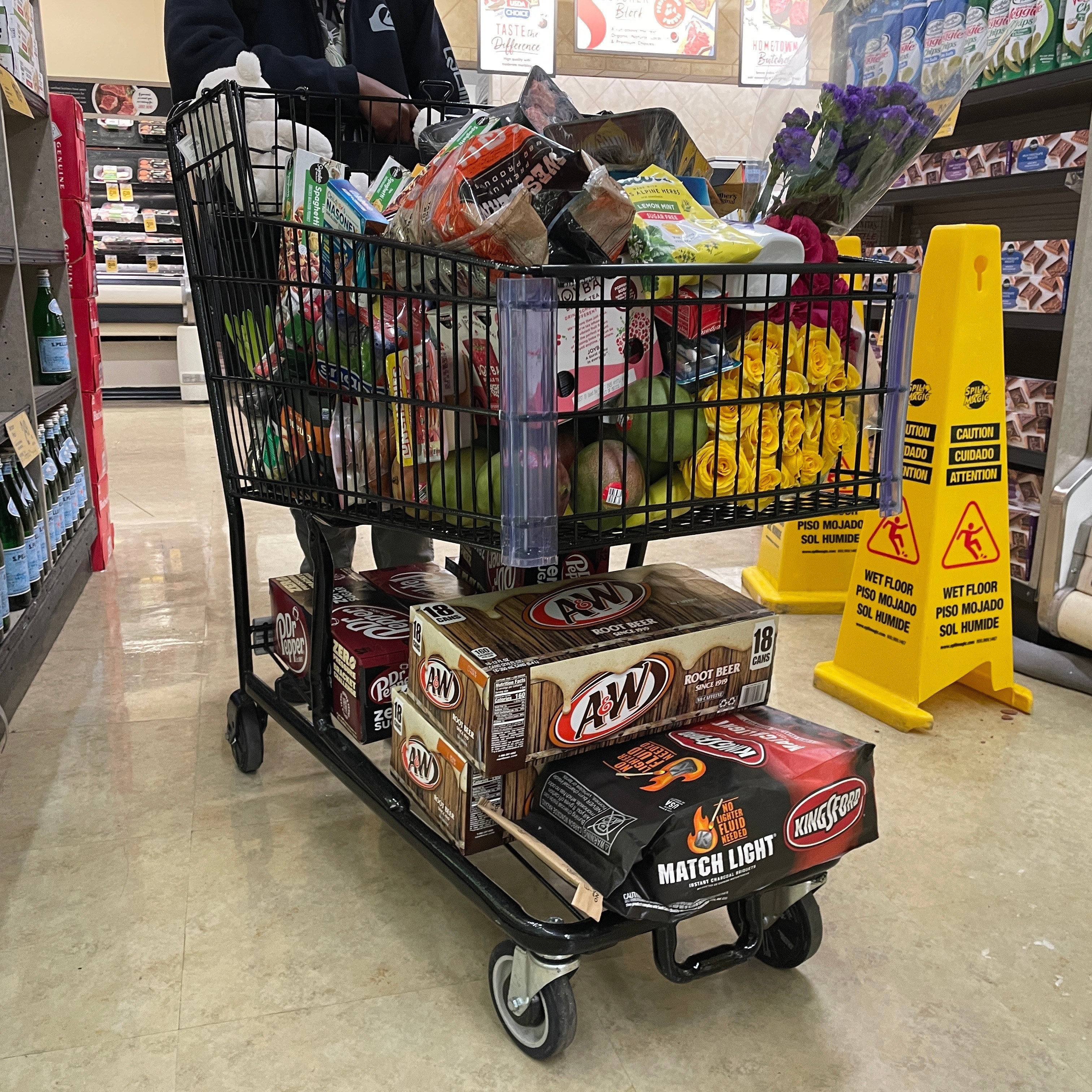 A shopping cart is filled to the brim with groceries, including flowers. Two boxes of root beer and charcoal are beneath the cart. Caution signs are nearby.