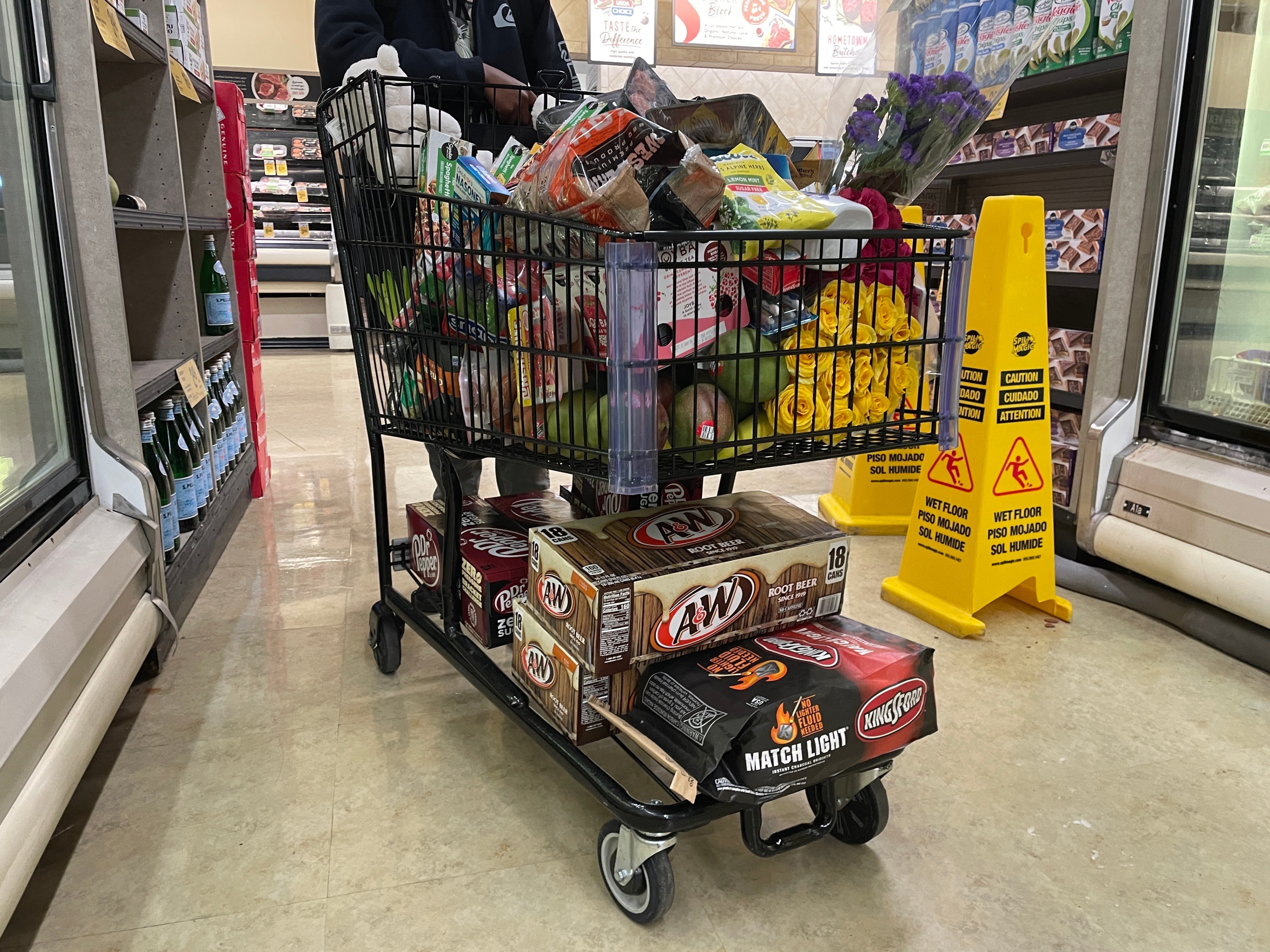 A shopping cart is filled to the brim with groceries, including flowers. Two boxes of root beer and charcoal are beneath the cart. Caution signs are nearby.