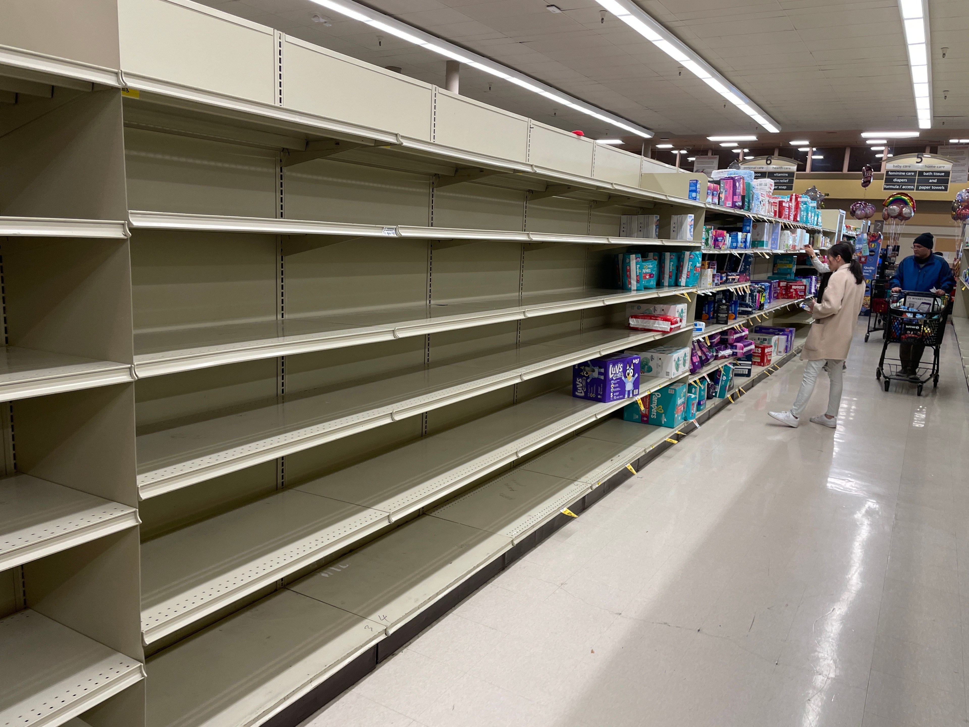 The image shows mostly empty store shelves on the left, with a few stocked shelves on the right. A person is shopping and another person pushes a cart.