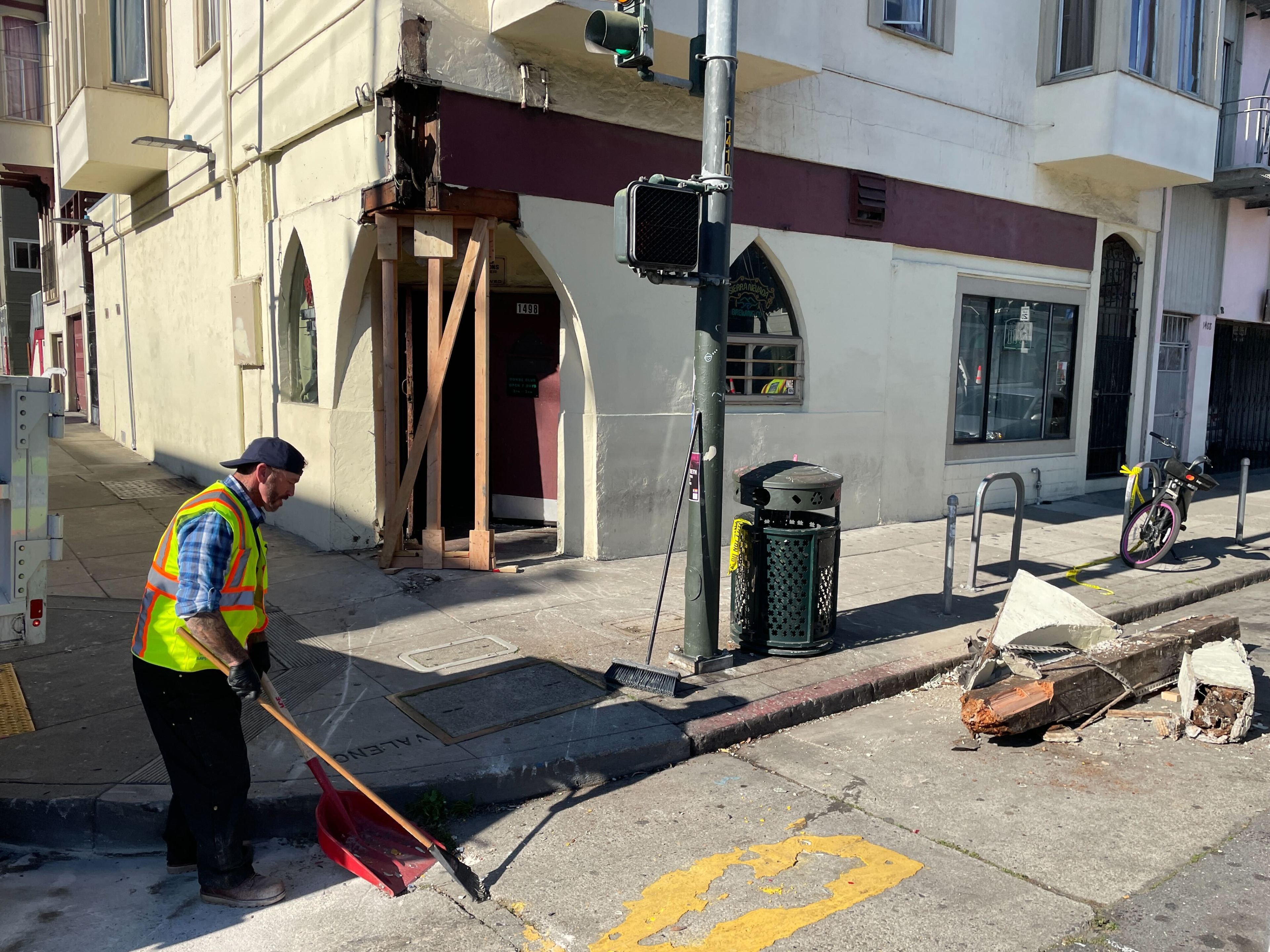 A worker in a high-visibility vest sweeps debris on a sunlit city sidewalk by a damaged corner building with wooden supports and a parked bicycle nearby.