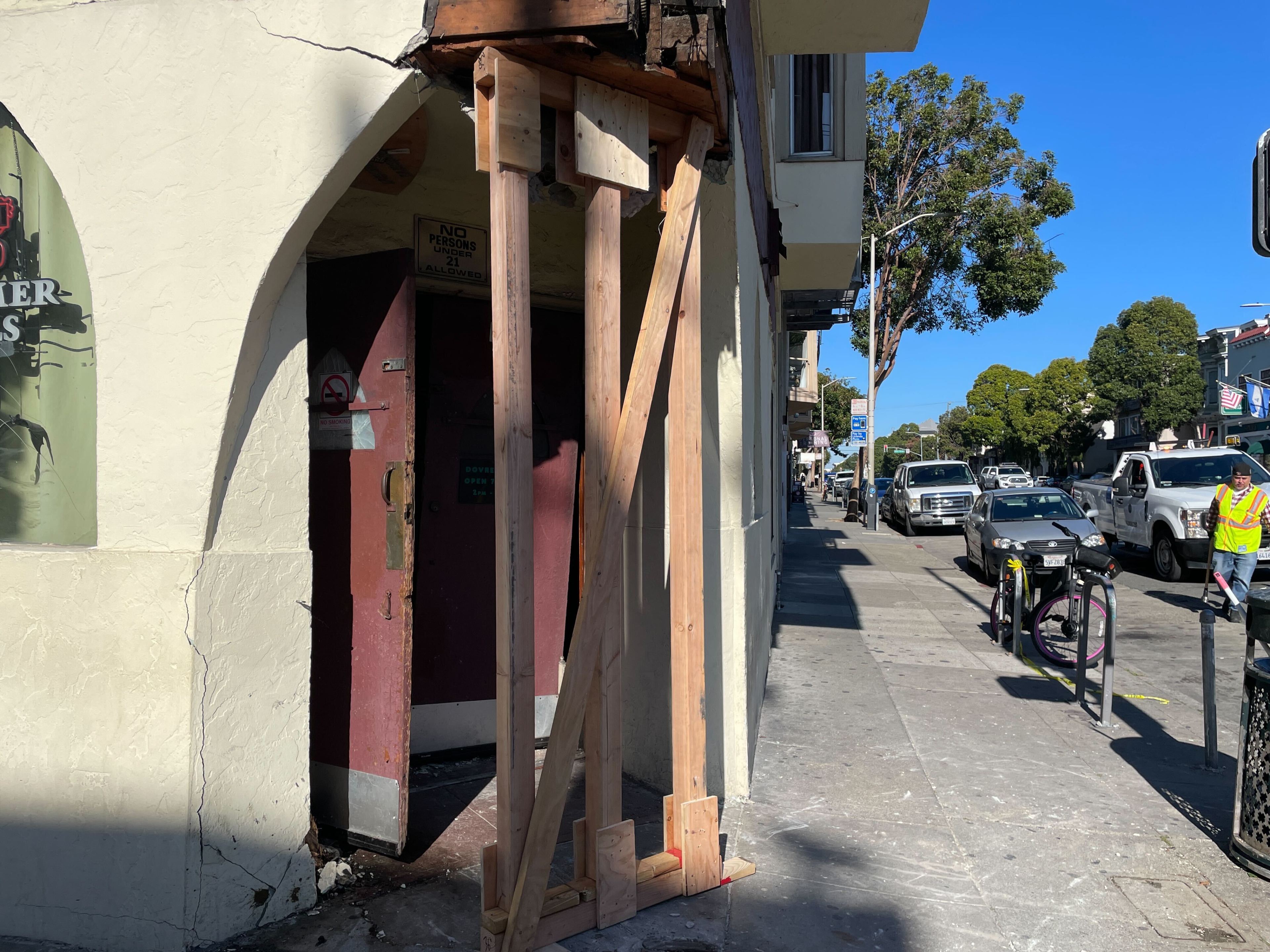 A damaged building corner supported by wooden beams is seen. A sign reads &quot;No persons under 21 allowed.&quot; Nearby, cars and a person in a safety vest are on the street.