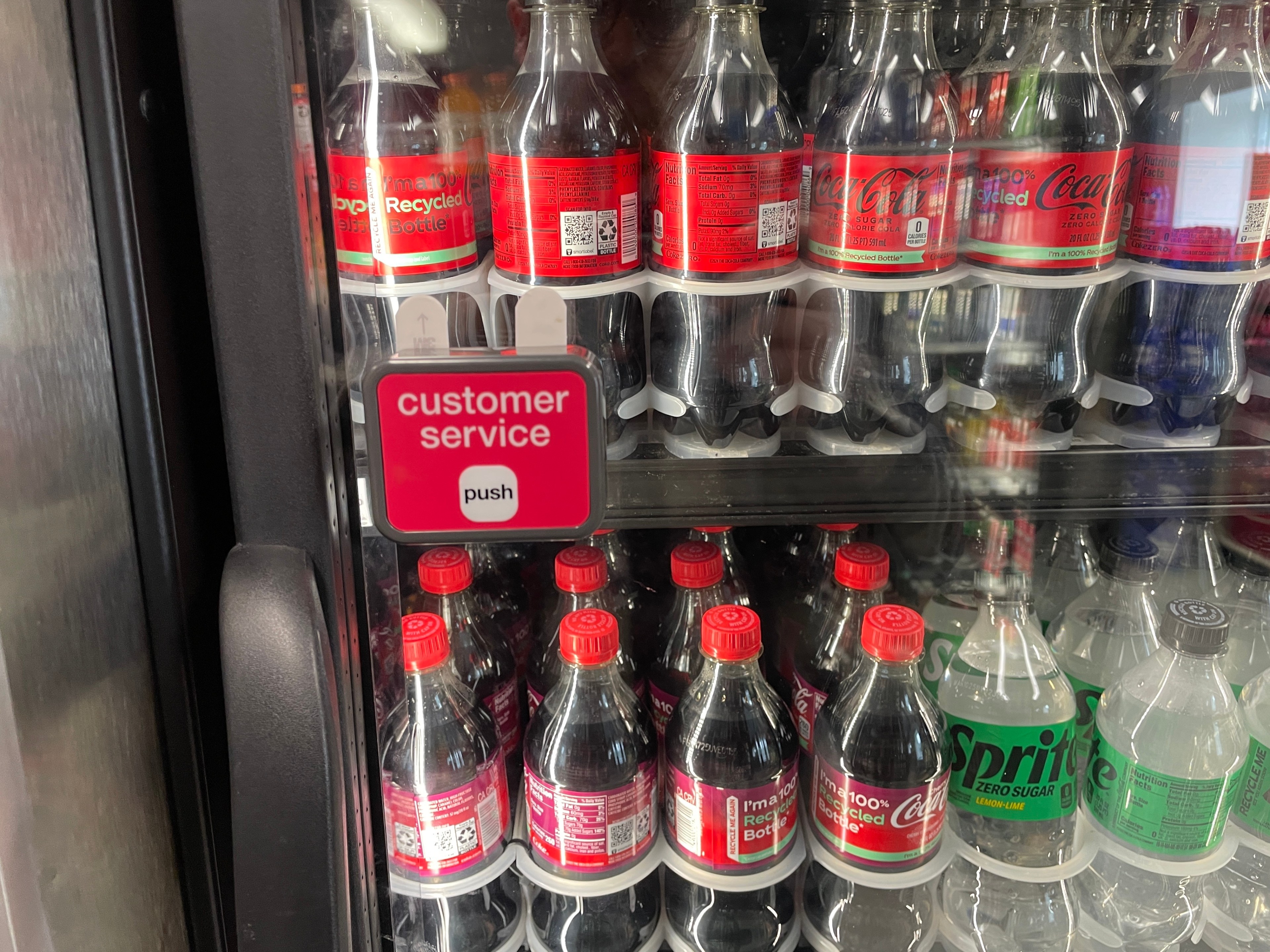 A cooler showcases rows of Coca-Cola and Sprite bottles. A &quot;customer service&quot; button labeled &quot;push&quot; is attached to the cooler door.