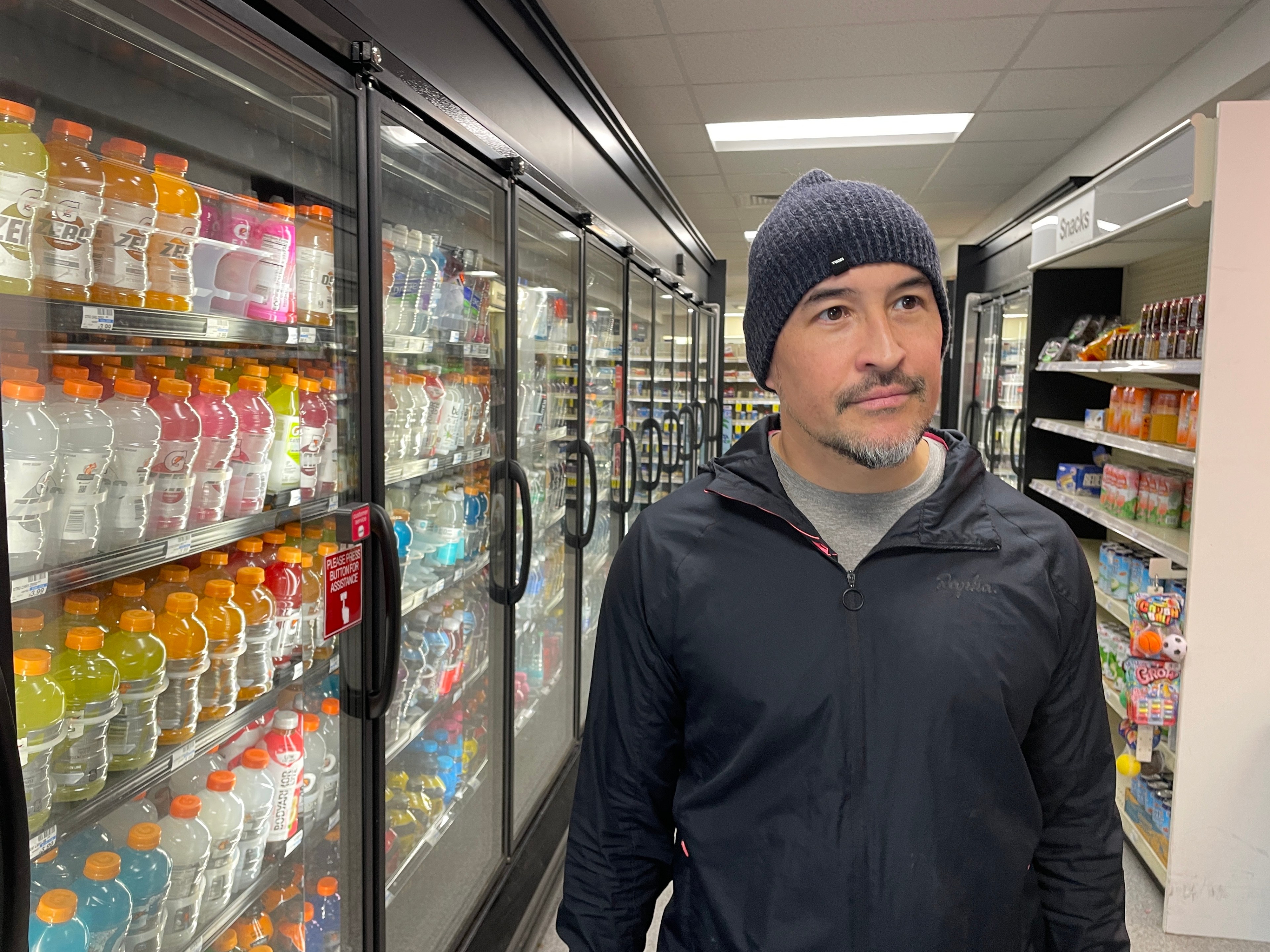 A man in a black jacket and beanie stands in a store aisle with various drinks in refrigerated cases on the left and snacks on the right.