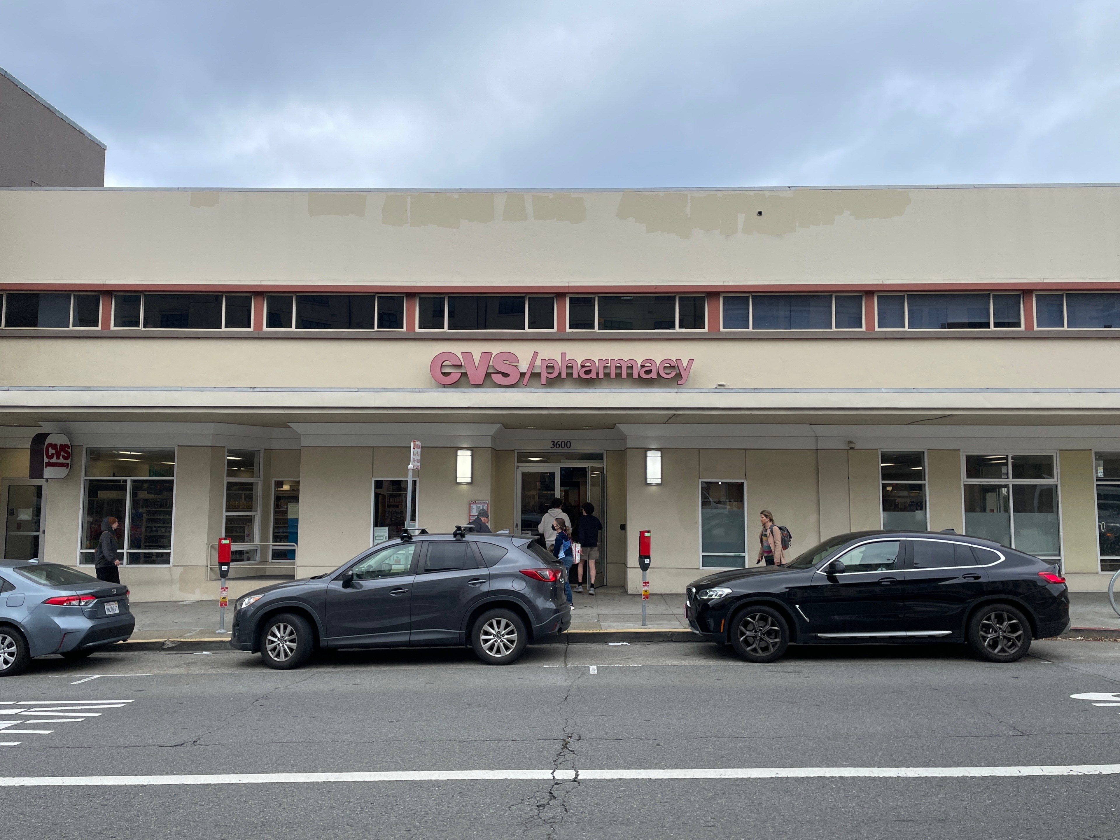 The image shows a CVS Pharmacy with a beige facade. Two cars are parked in front, and several people are entering or exiting the store.