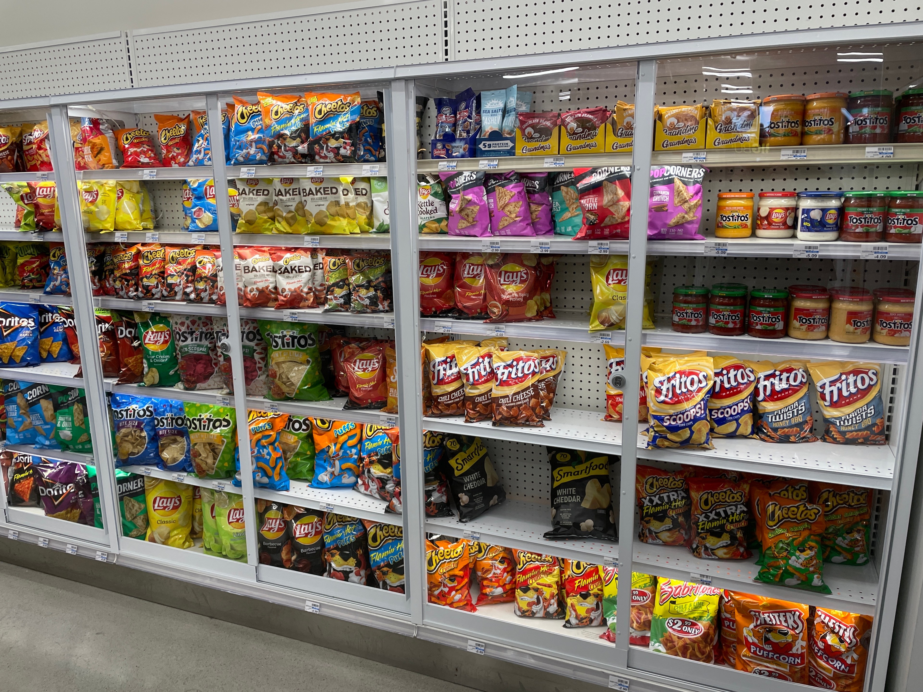 The image shows shelves in a store stocked with various chips and snacks, including brands like Cheetos, Fritos, Lays, Tostitos, and popcorn.