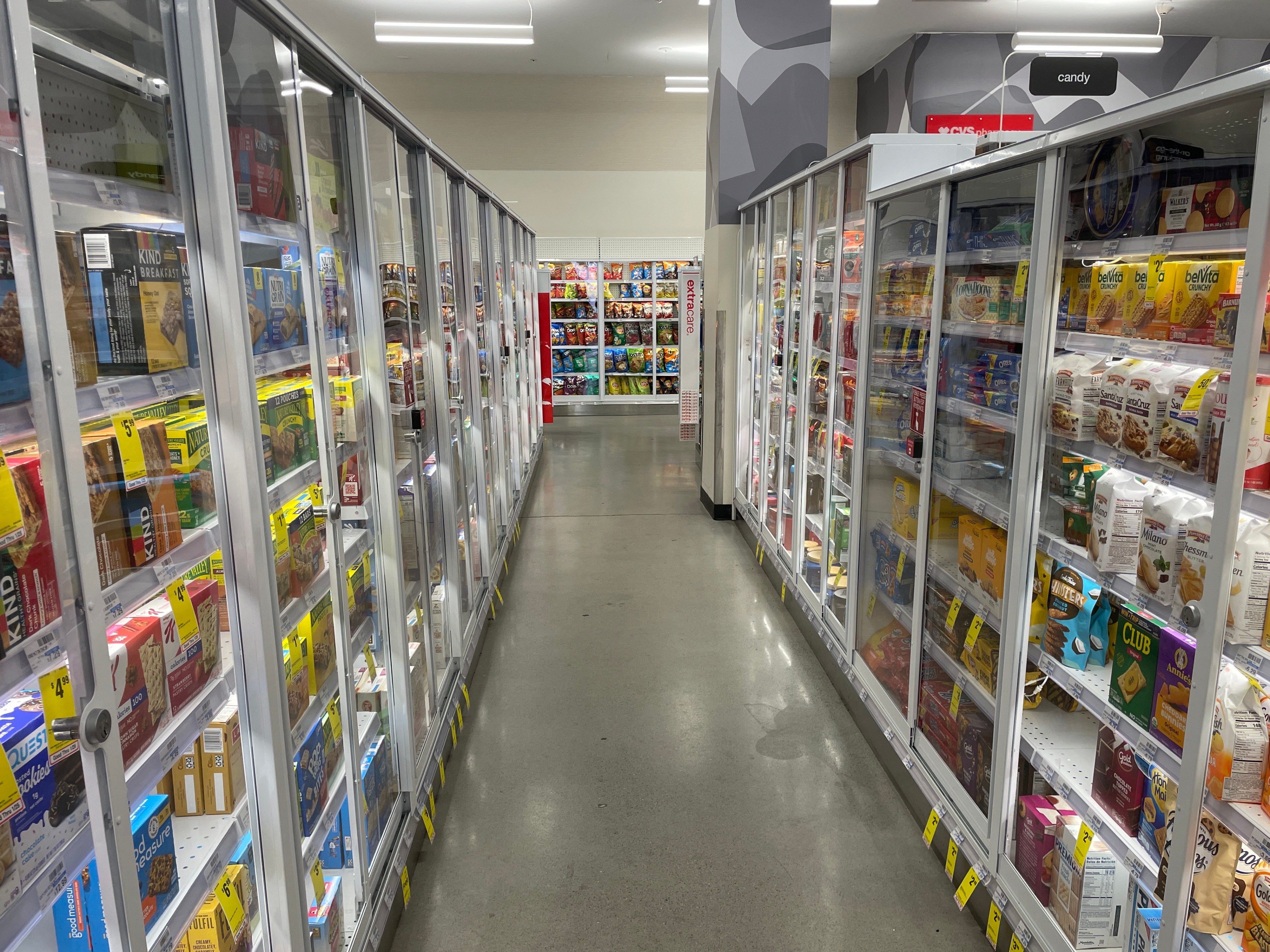 The image shows an aisle in a store with glass-door freezers on both sides filled with snacks and packaged food. The floor is smooth and polished.