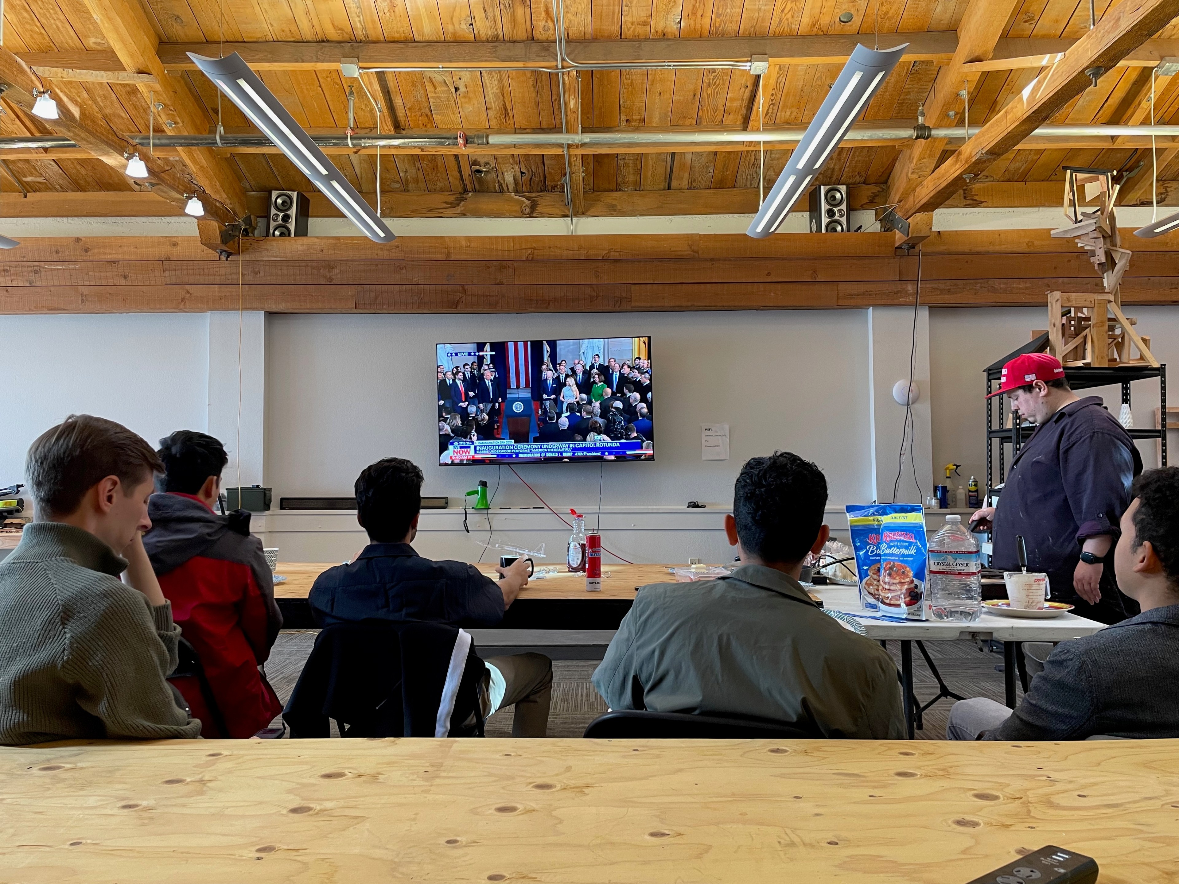 A group of people in a room with wooden beams watch a news event on a wall-mounted TV. Pancake mix and syrup are on a table with other items.