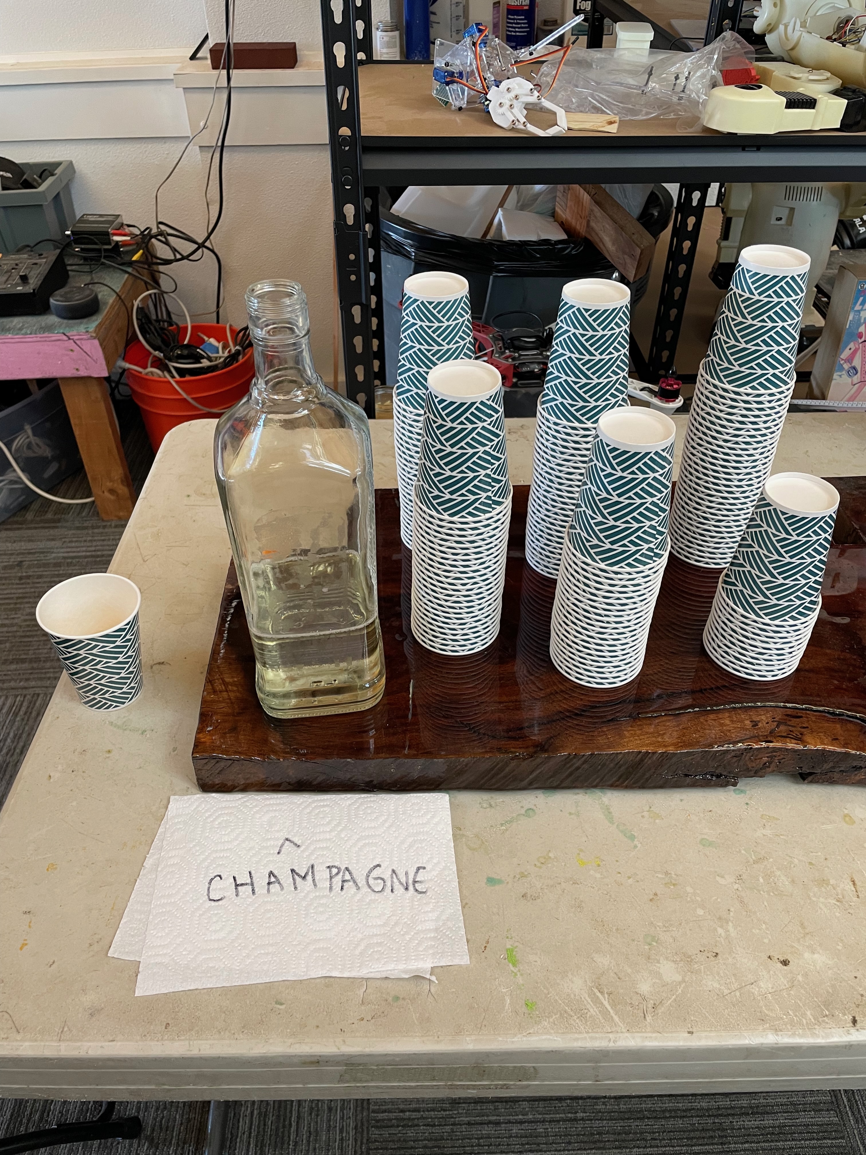 Cups are arranged on a wood cutting board, alongside a glass bottle full of liquid. A sign written on a napkin, pointing toward the bottle, reads, &quot;champagne.&quot;