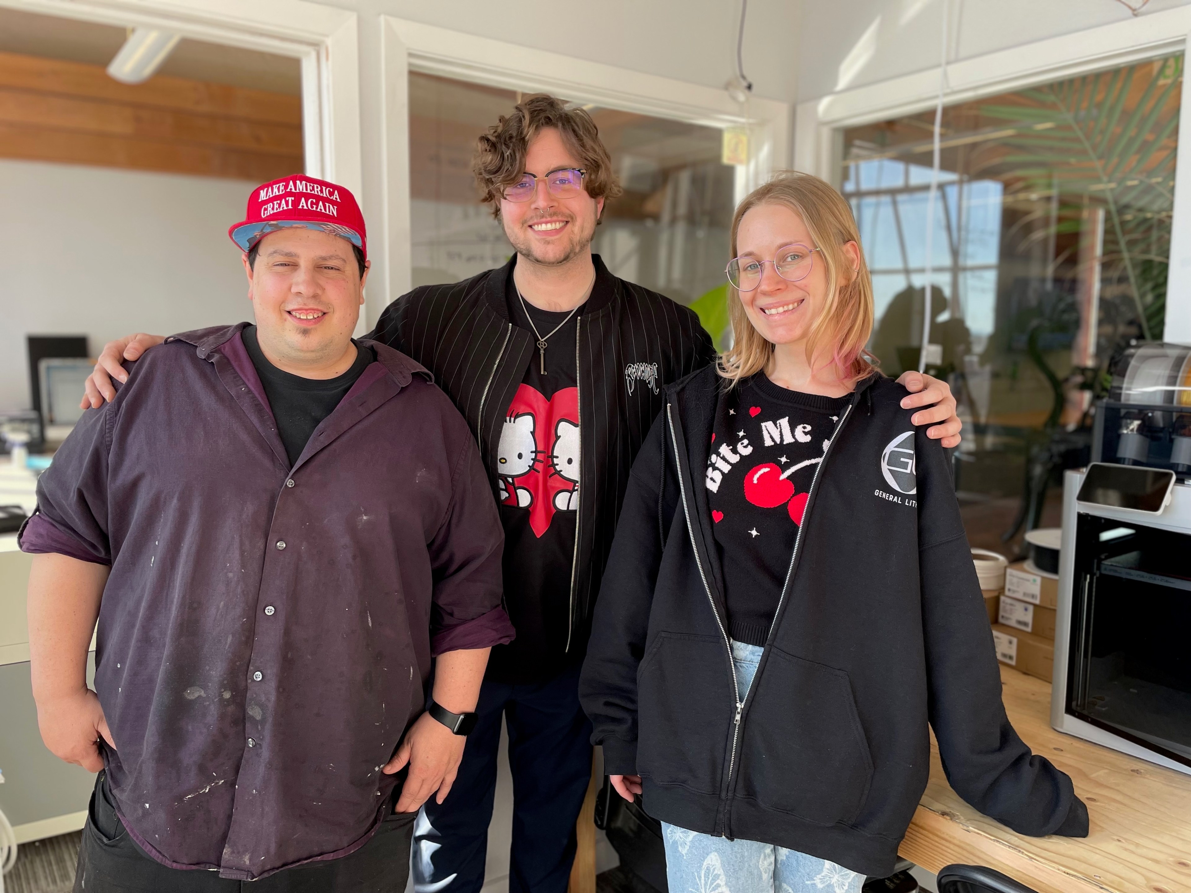 Three people are smiling in an office. One wears a red hat, another has a cartoon shirt, and the third is wearing glasses and a hoodie.