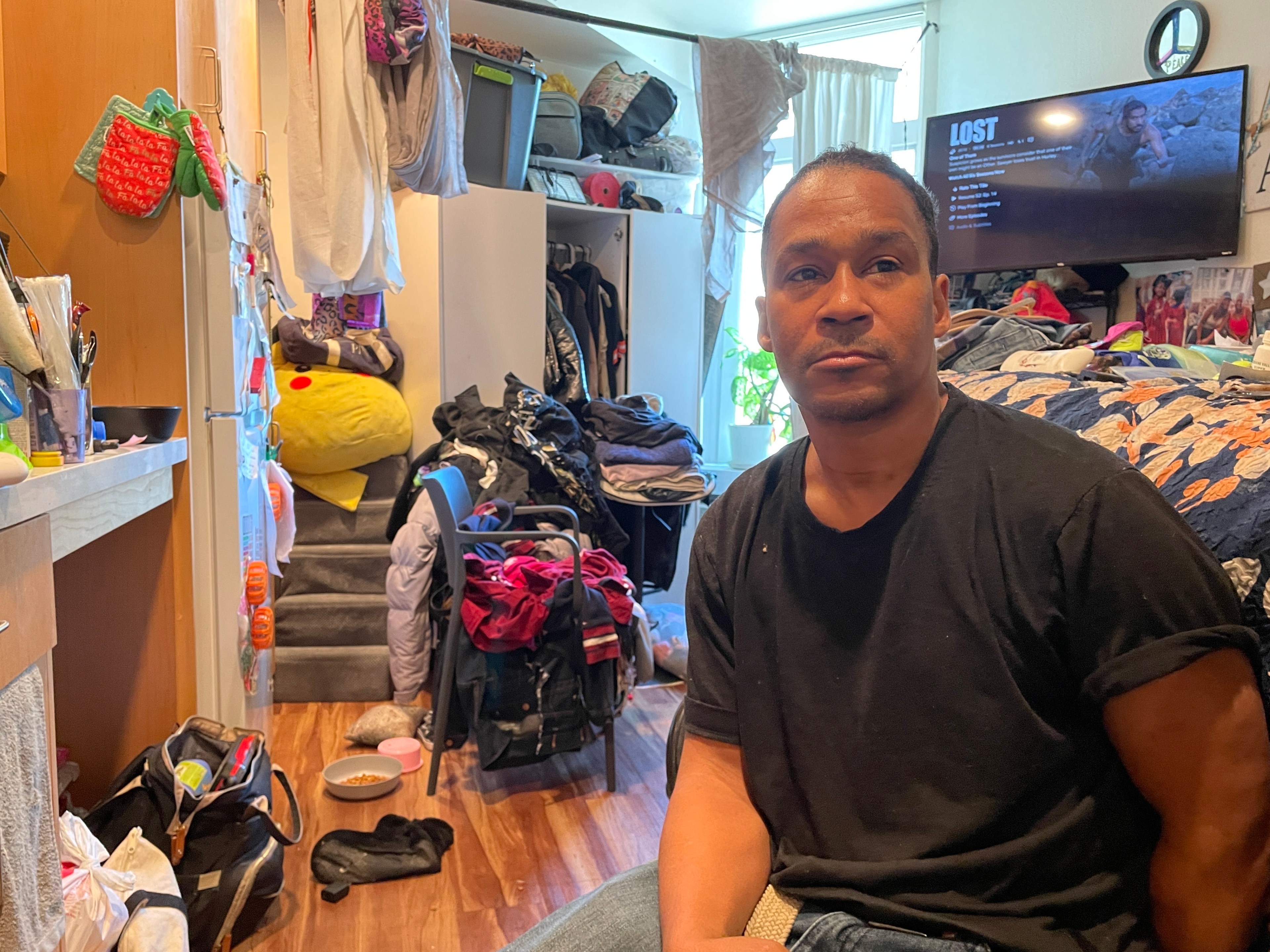 A person sits in a cluttered room with clothes and items scattered around. A TV screen is on, showing a scene from &quot;Lost.&quot; Natural light streams in from a window.