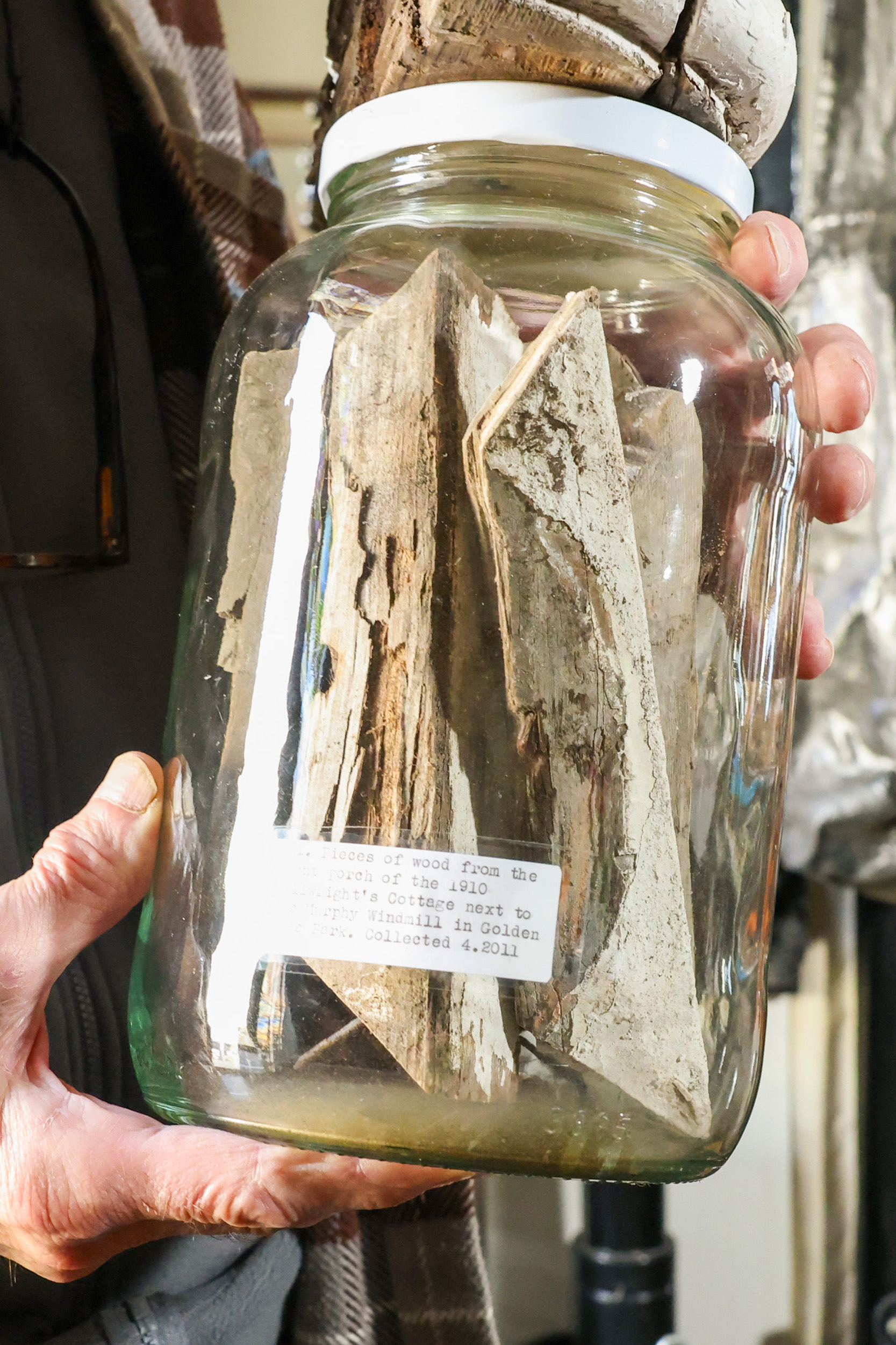 A hand holds a glass jar with pieces of aged wood inside. A label on the jar describes the wood's historical significance and collection date.