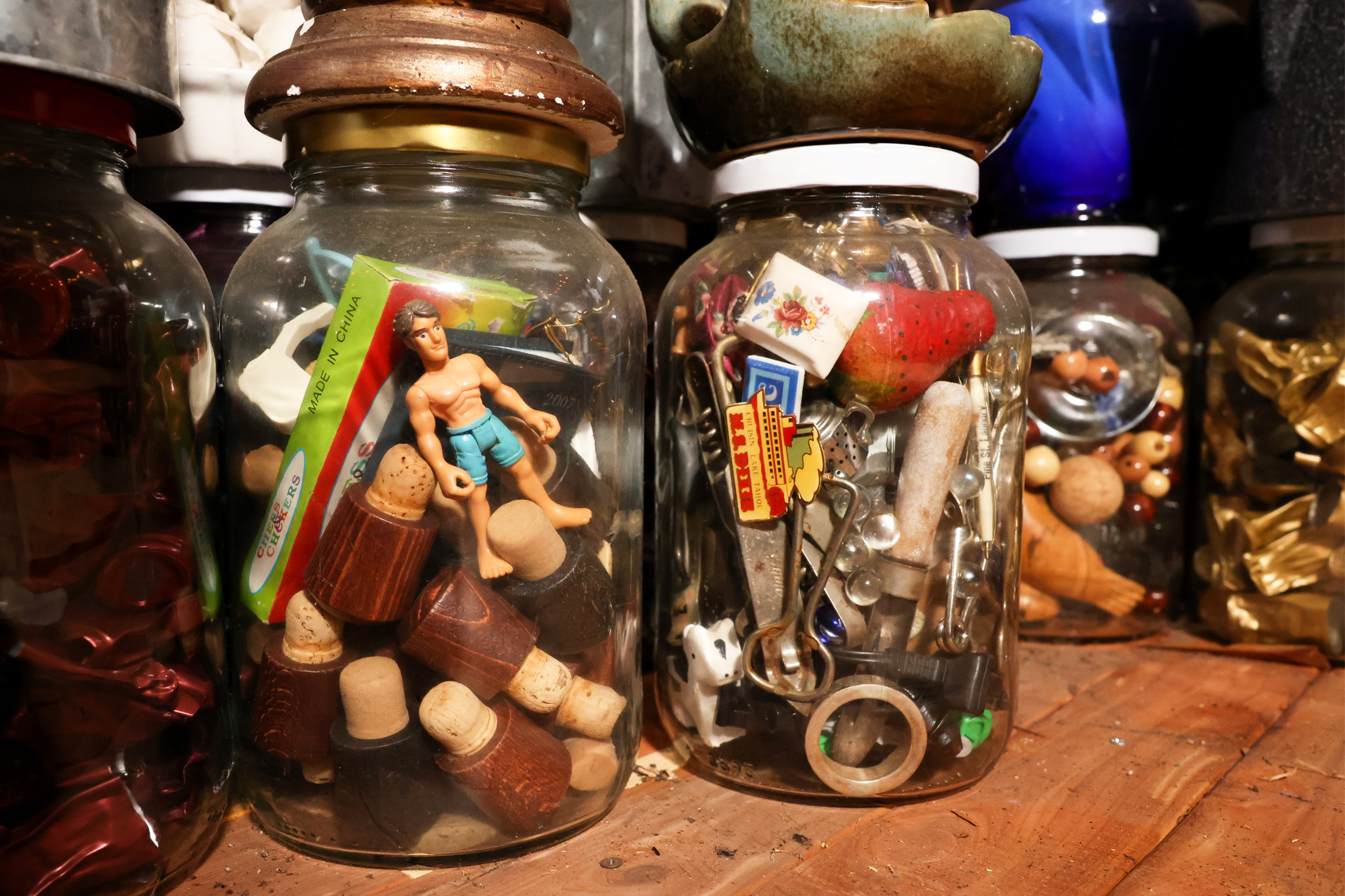 The image shows jars filled with assorted items: a toy figure, corks, small objects like keys and buttons, set on a wooden surface.