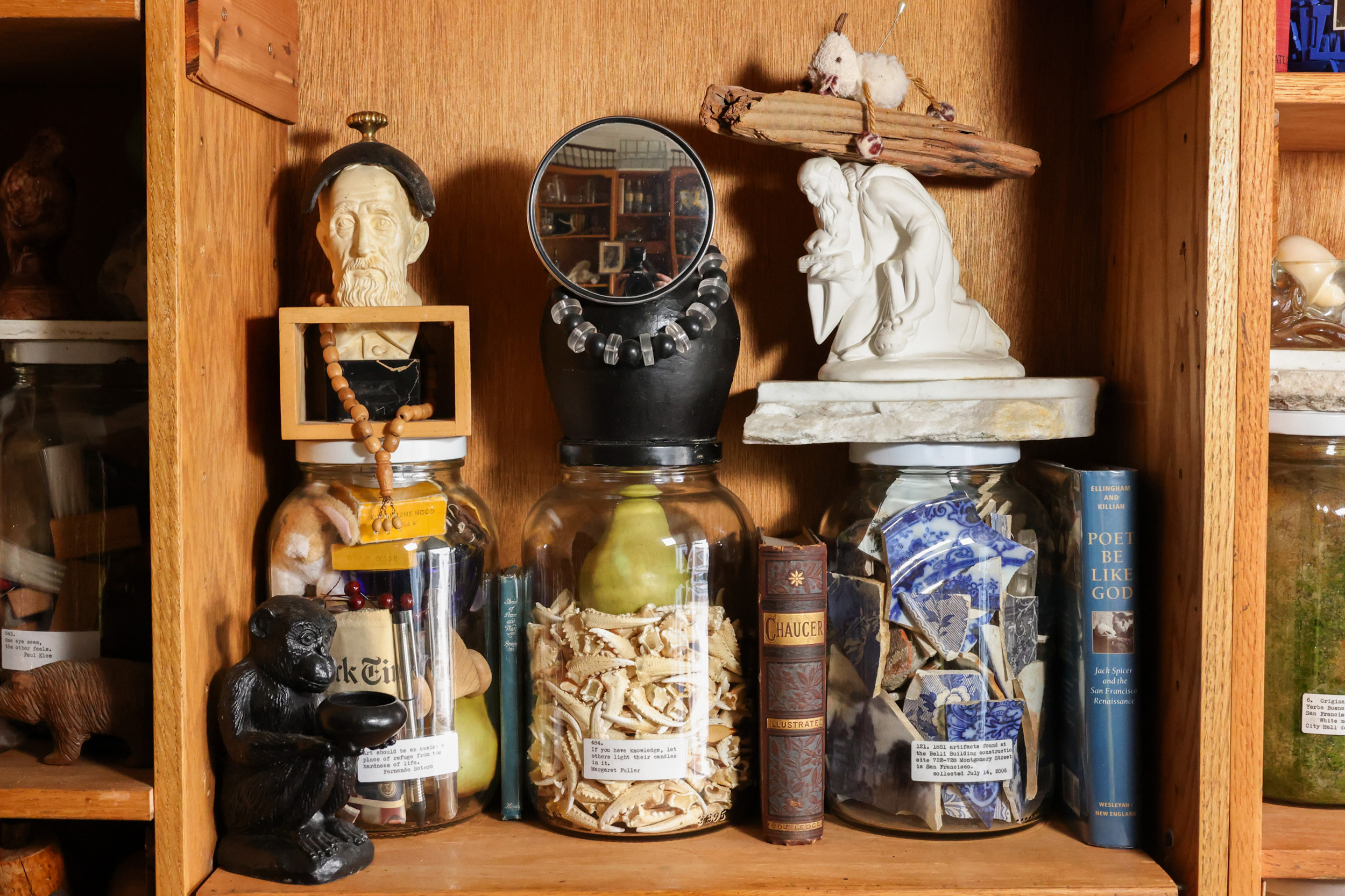 The image shows a shelf with eclectic items: jars contain bones, blue pottery shards, and more. Books and sculptures, including a bust and figure, add to the collection.