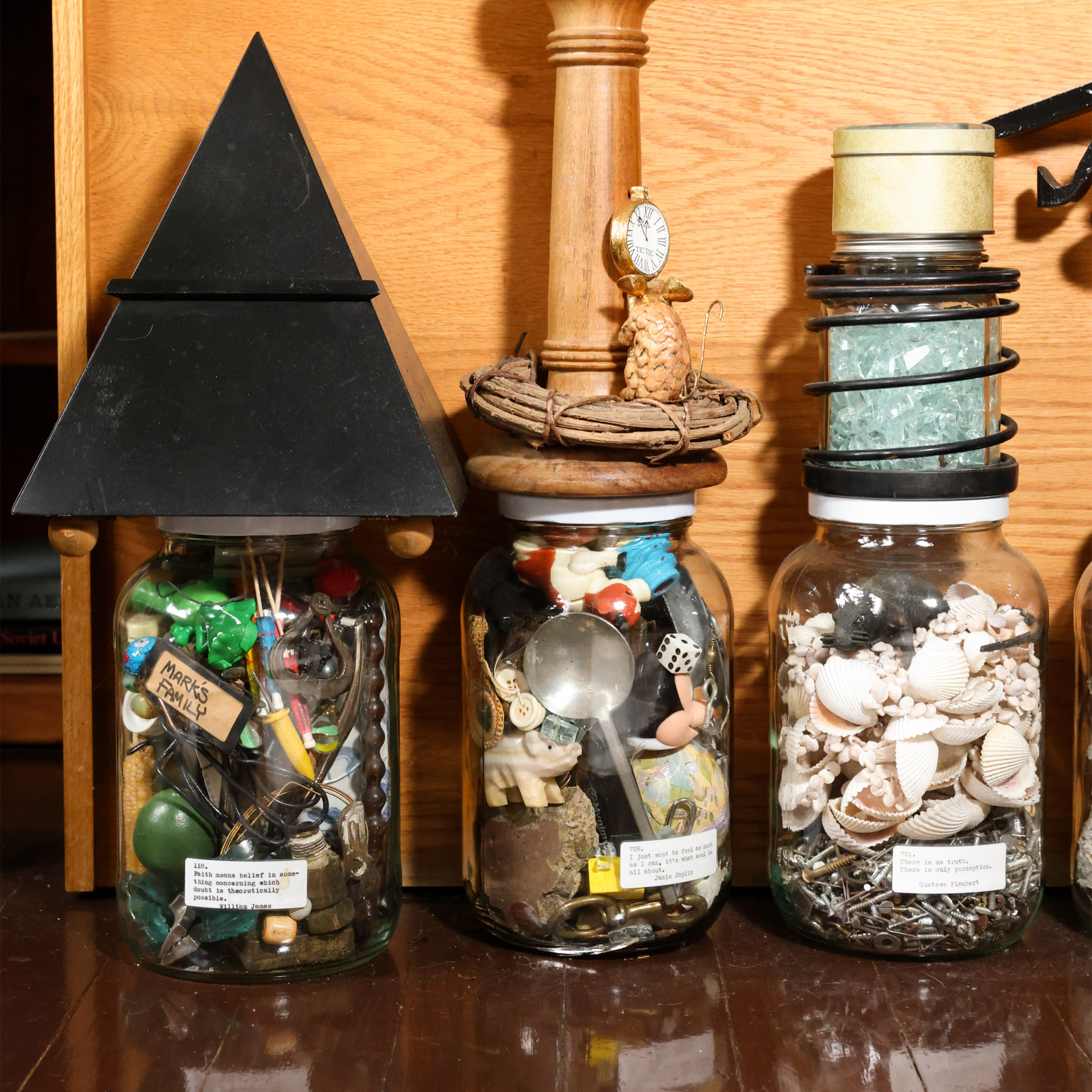 Three glass jars filled with assorted objects sit on a wooden shelf. The left jar has a black pyramid lid, the middle features a small clock, and the right jar is topped with a coil.