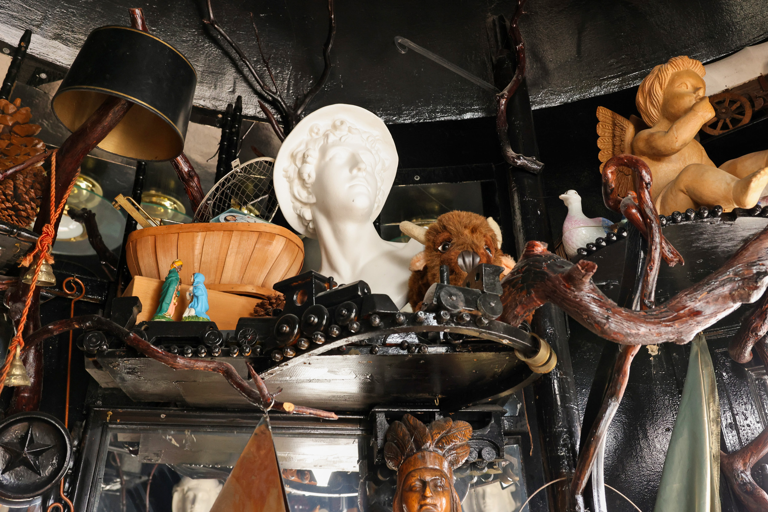 The image features a cluttered display with a white bust, a wooden bowl, toy figures, a buffalo plush, and a small cherub statue amidst branches and eclectic decor.