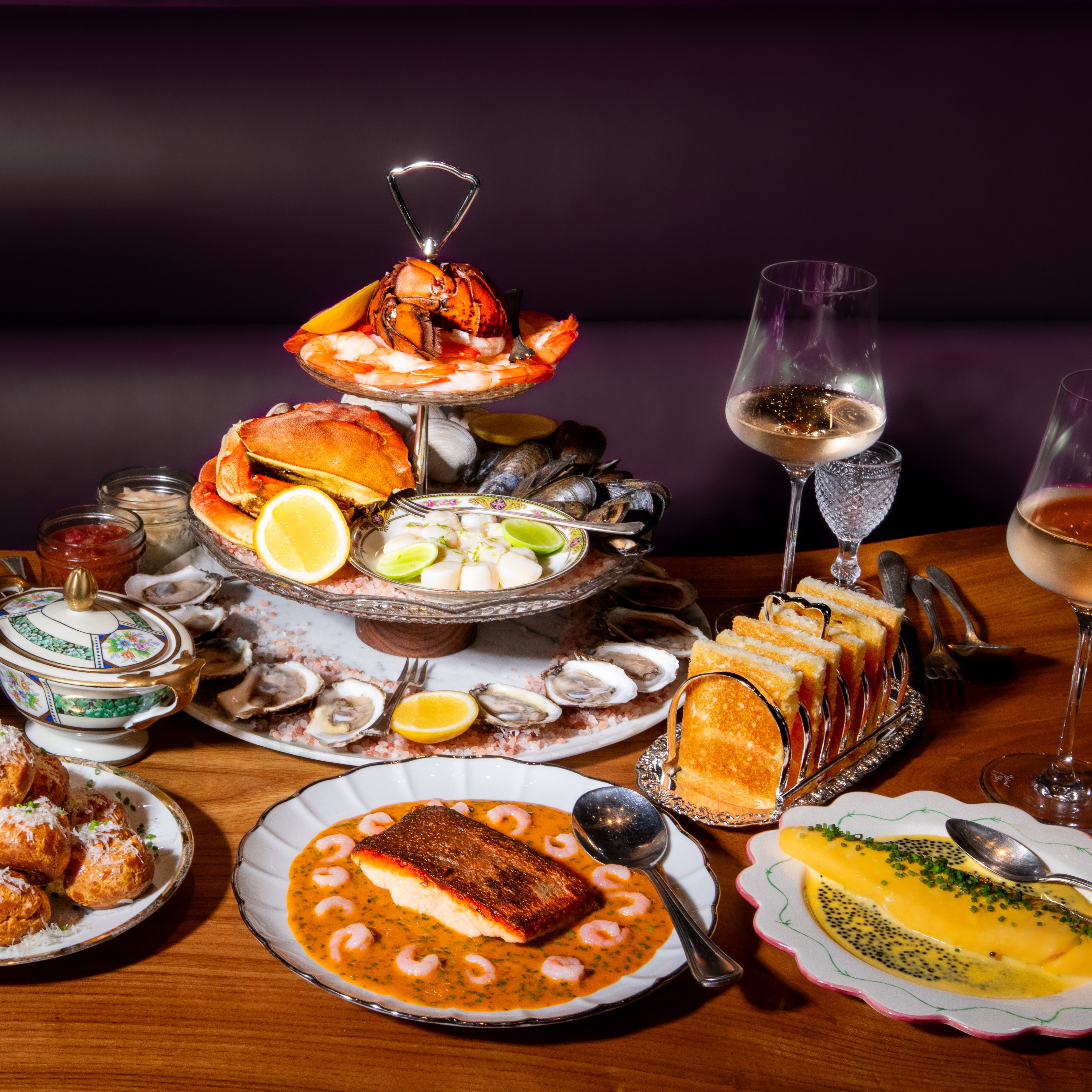 A table is set with a seafood tower, salad, fries, toasted bread, salmon with sauce, and assorted pastries. Four glasses of white wine accompany the dishes.