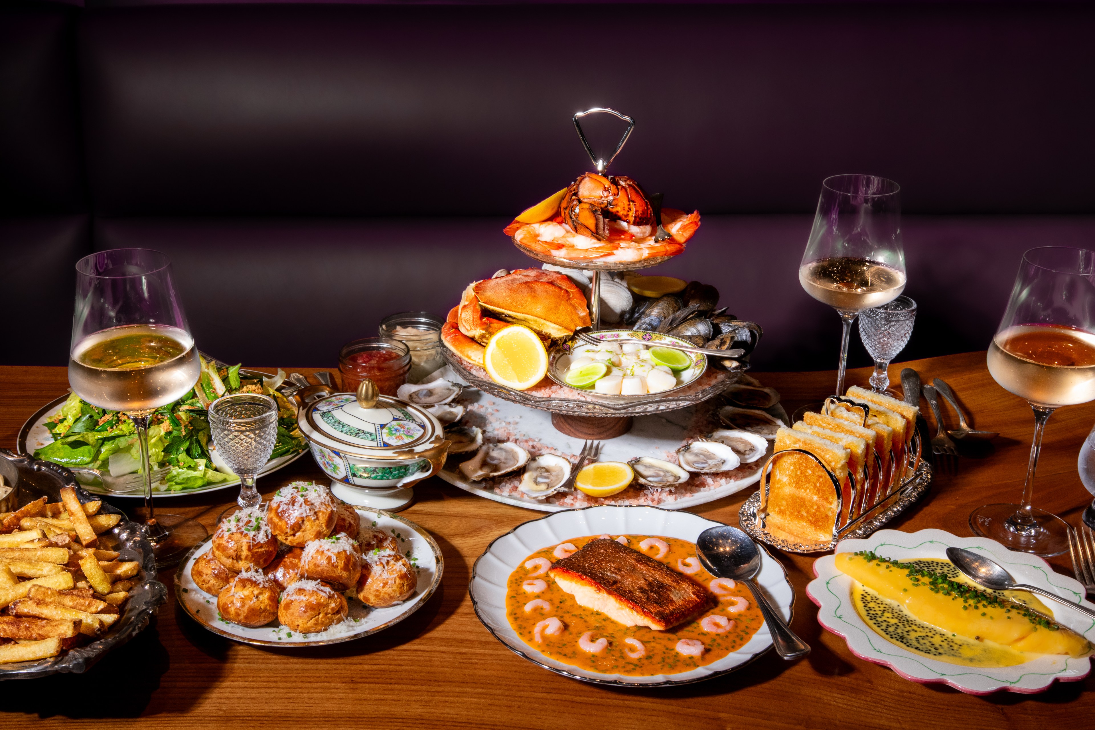 A table is set with a seafood tower, salad, fries, toasted bread, salmon with sauce, and assorted pastries. Four glasses of white wine accompany the dishes.