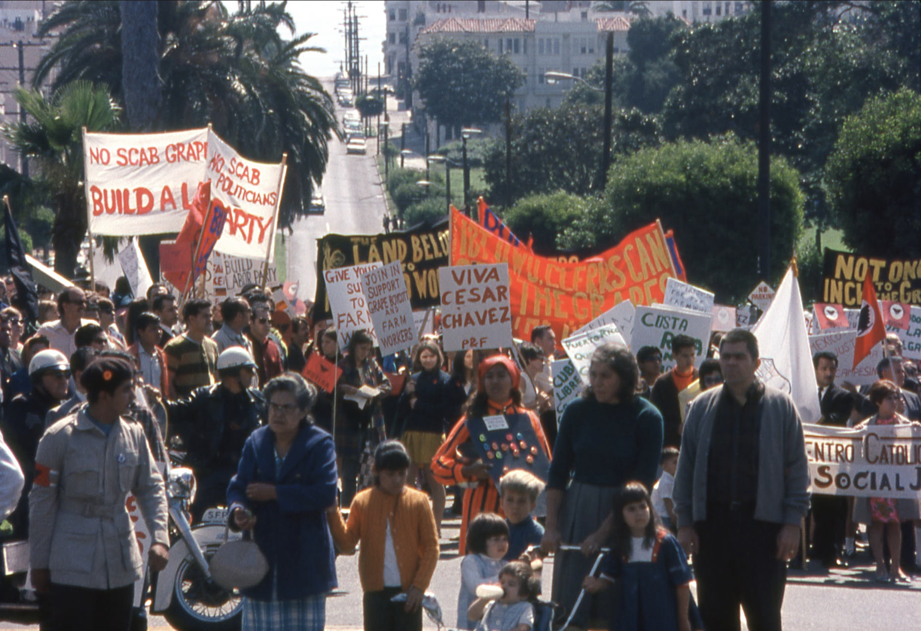 A large group of people are marching with colorful signs and banners advocating for workers' rights and supporting Cesar Chavez. Police officers on motorcycles are present.