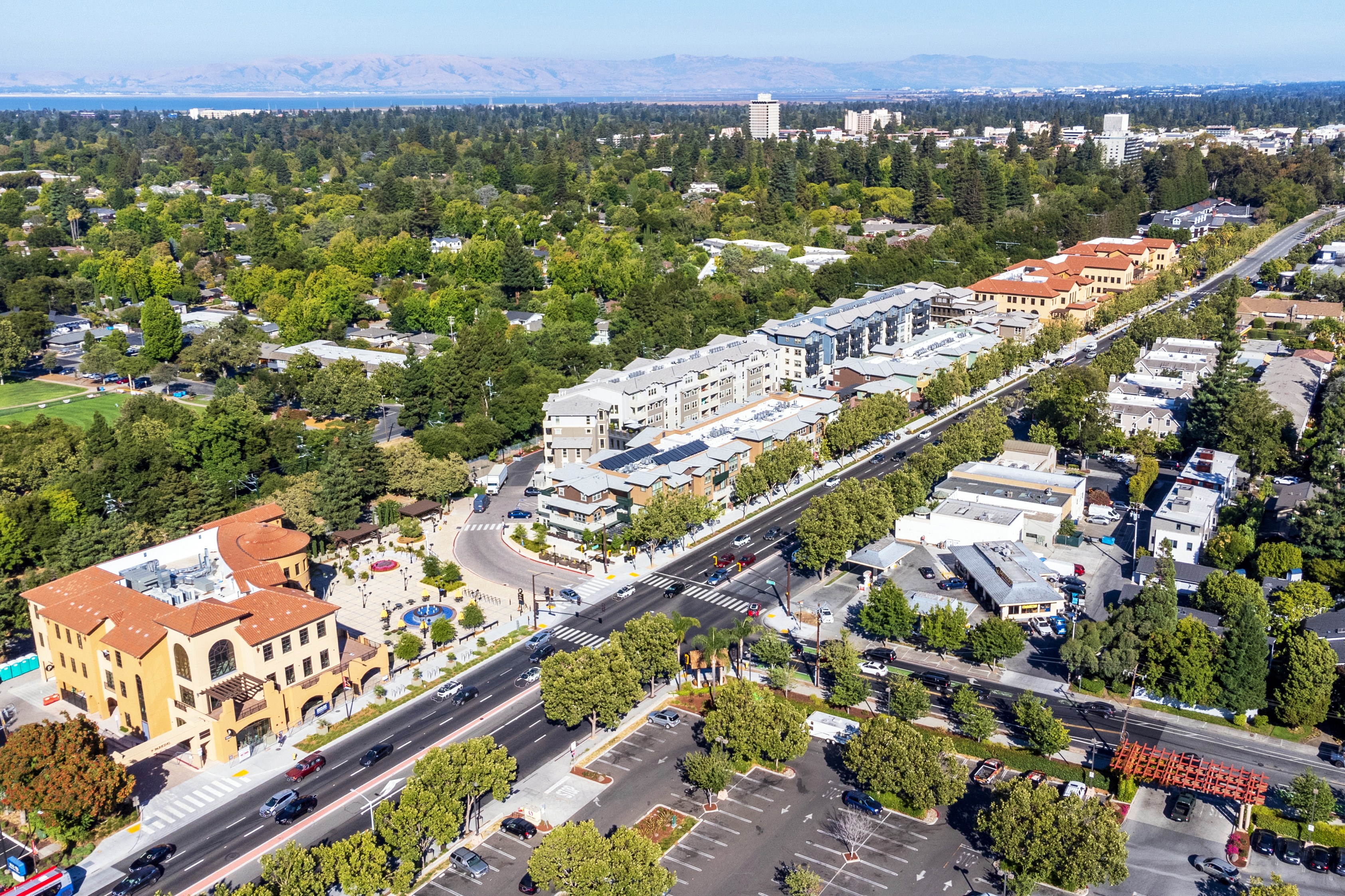 The image shows an aerial view of a suburban area with tree-lined streets, residential buildings, a baseball field, and some parking lots, under a clear sky.