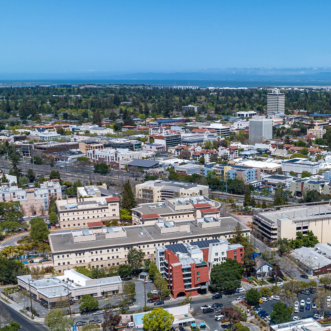 The image shows a cityscape with a mix of low-rise and mid-rise buildings surrounded by trees. In the background, there are distant hills under a clear blue sky.