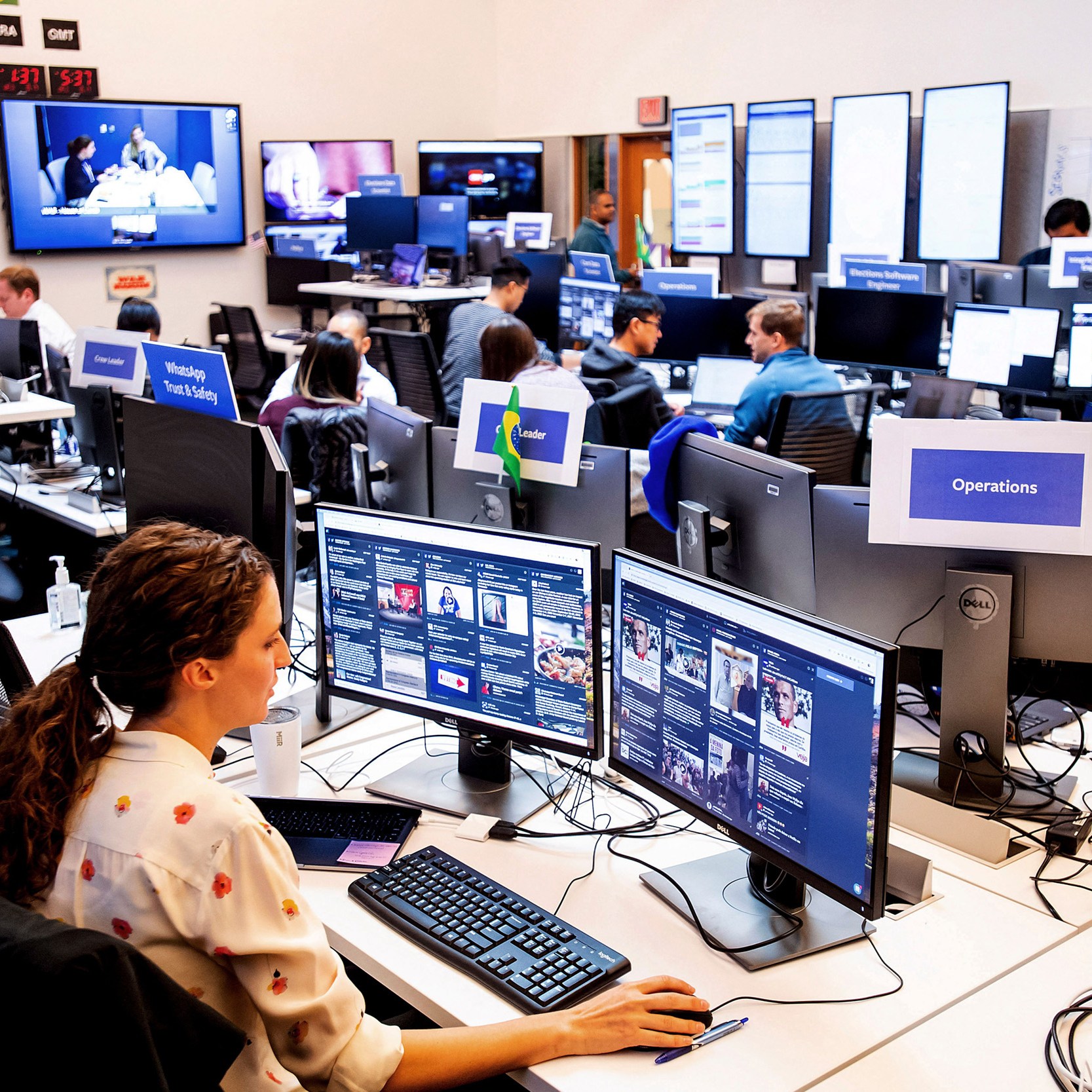 People work at a busy control center with numerous monitors, displaying various digital interfaces and video feeds, under labeled sections like "Operations."