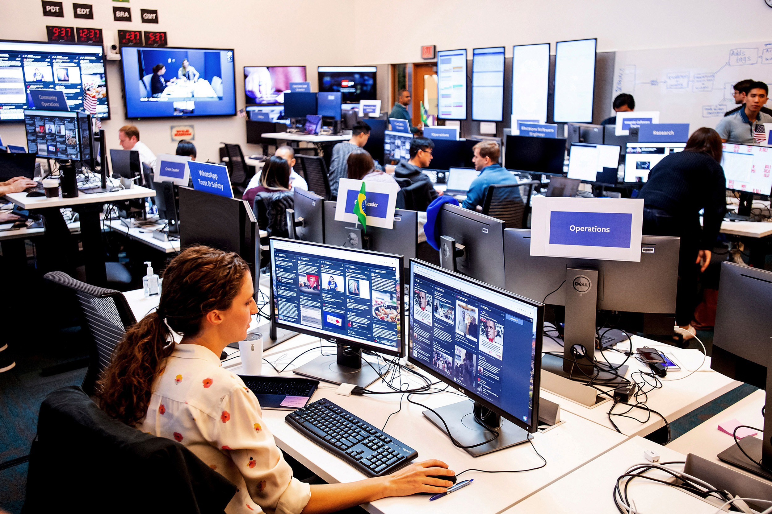 People work at a busy control center with numerous monitors, displaying various digital interfaces and video feeds, under labeled sections like "Operations."