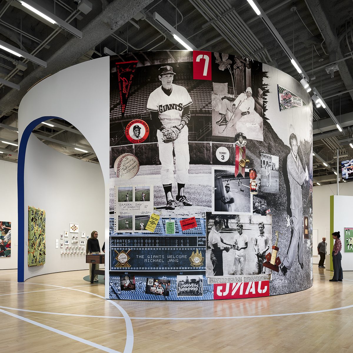 The image shows a curved wall in a gallery covered with vintage baseball photos and memorabilia. A woman stands nearby, and the space has an arched entrance.