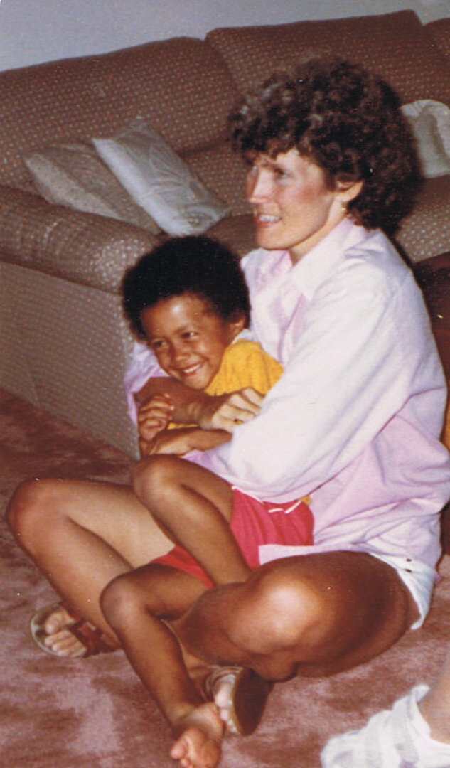 A smiling child in a yellow top and red shorts sits on a woman's lap. The woman has curly hair and wears a light pink shirt, sitting on a carpeted floor.