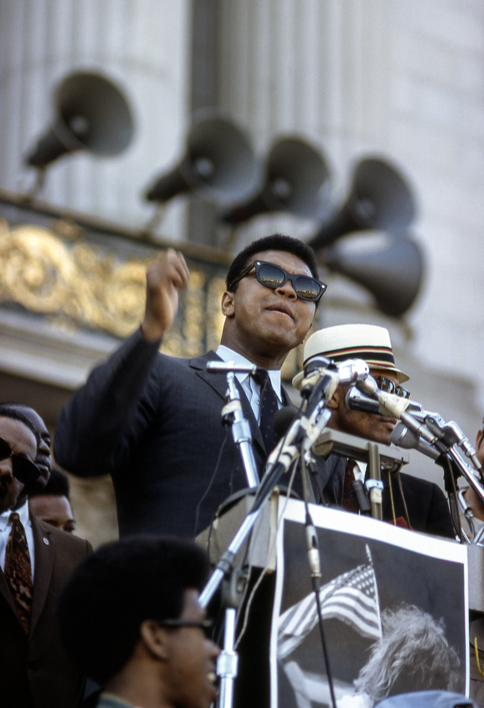 A man in a suit and sunglasses speaks passionately at a podium surrounded by microphones. Behind him, large megaphones are visible on a building facade.