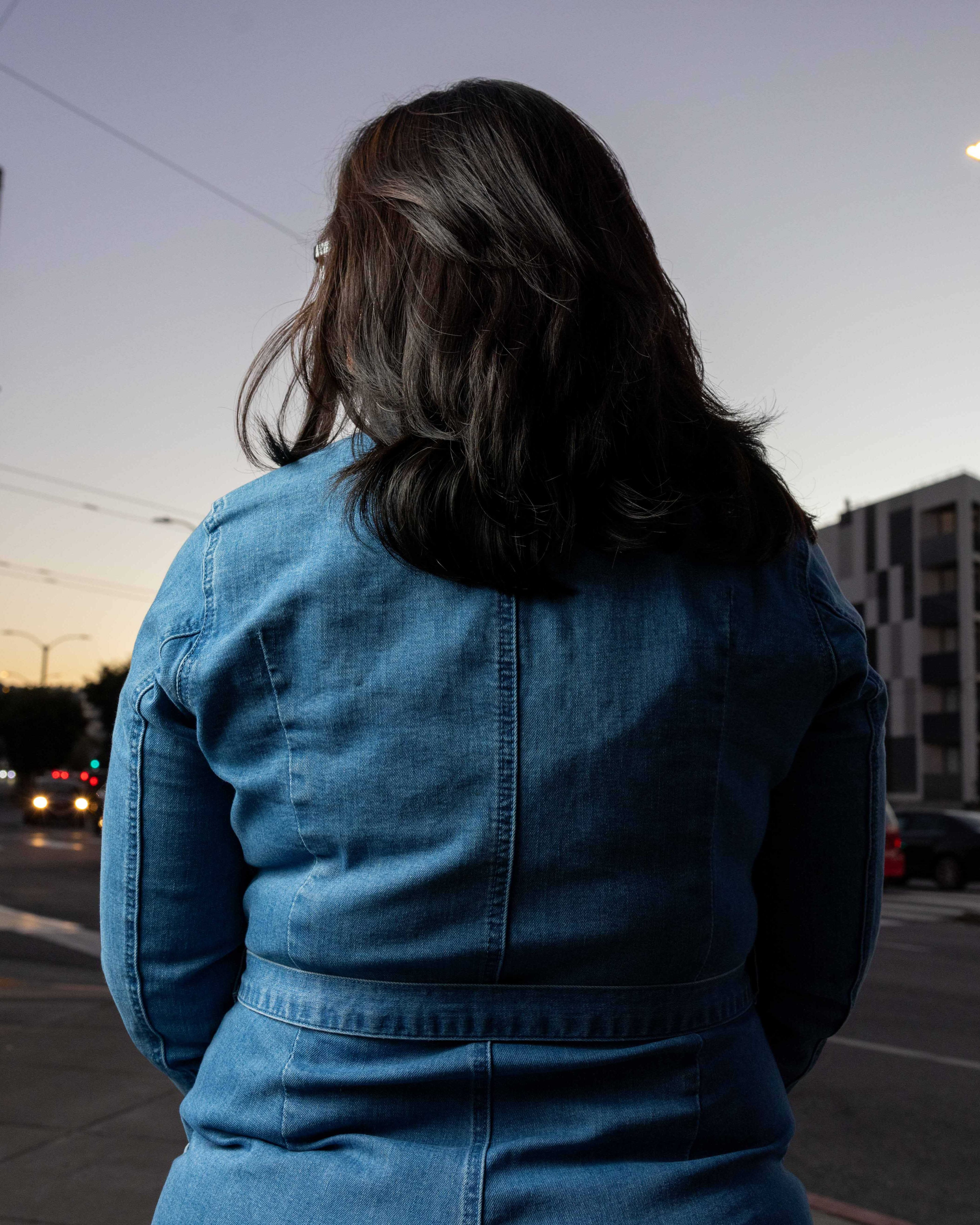 A person with long dark hair, wearing a blue denim jacket, stands facing away on a city street at dusk, with buildings and cars nearby.