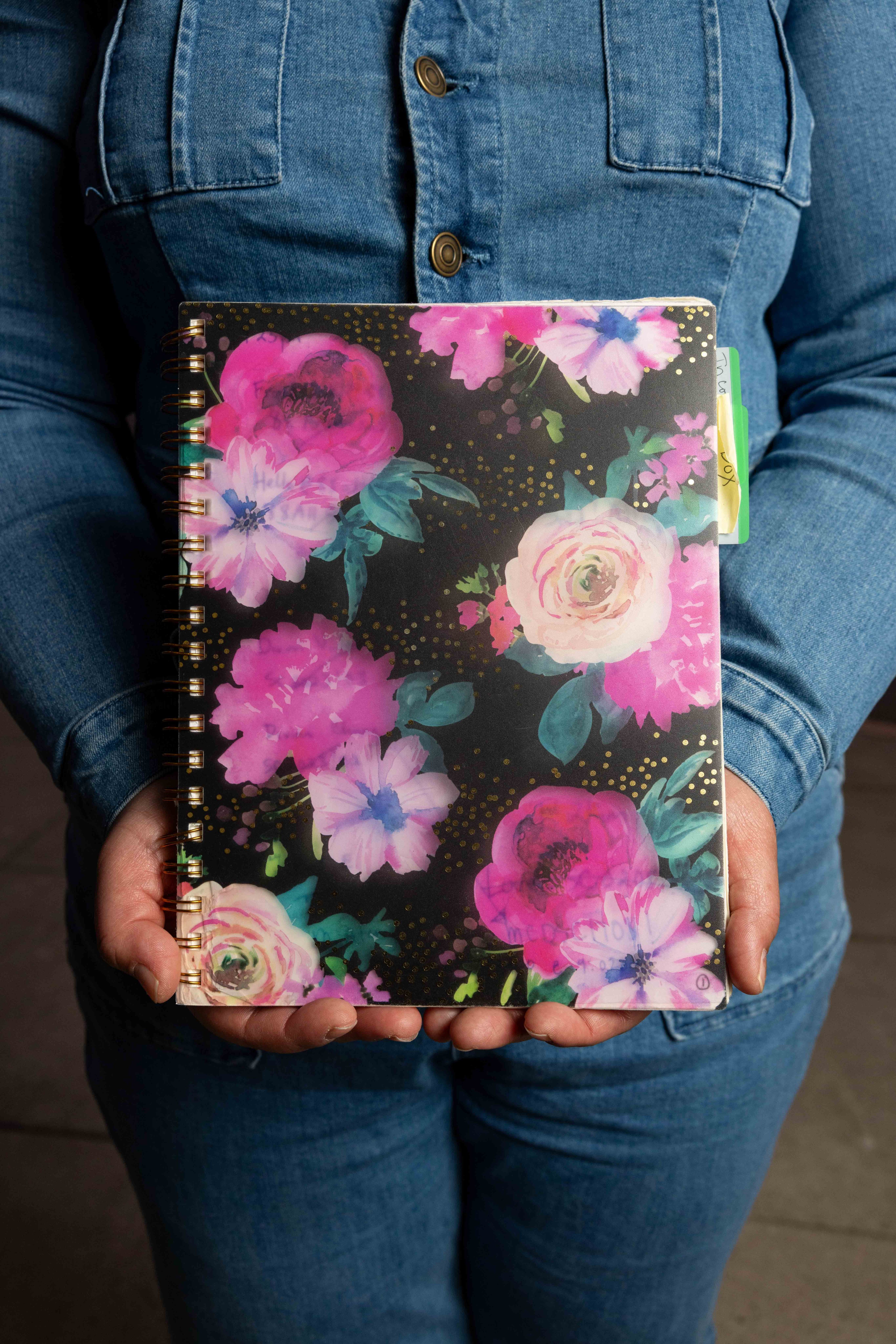 A person in a denim outfit holds a spiral notebook with a black cover, vibrant pink and purple floral design, and small gold dots.