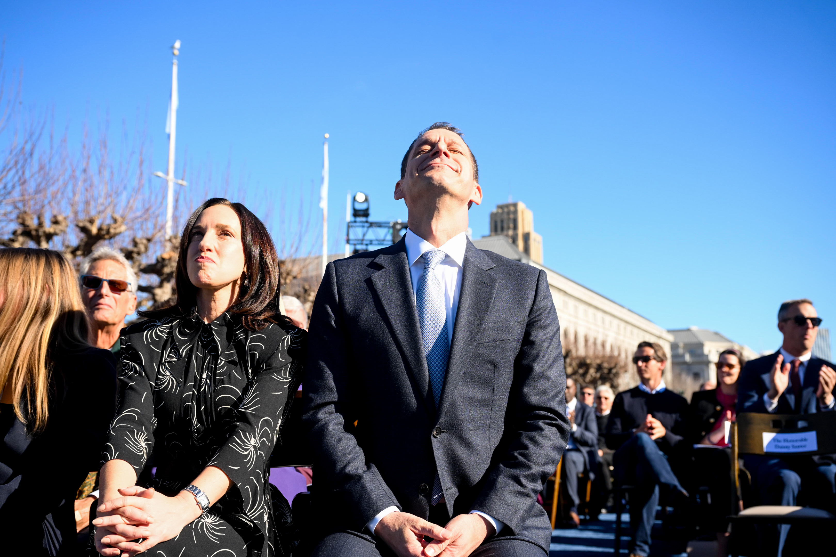 A group of people sit outdoors on a sunny day, some looking upward. A man in a suit in the center seems to be smiling with his eyes closed. A woman is sitting beside him.