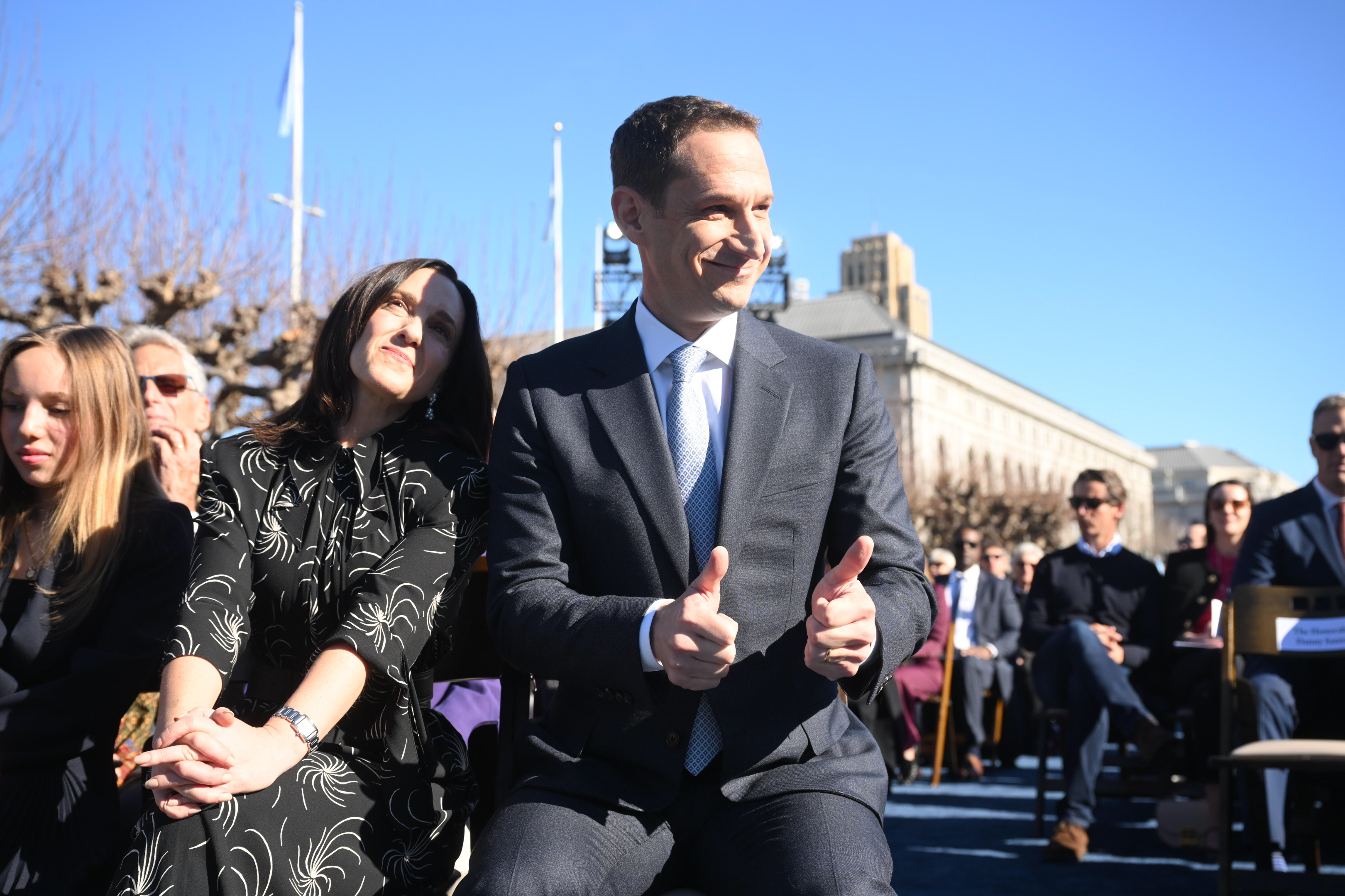 A man in a suit gives two thumbs up, and a woman next to him smiles, both seated outdoors on a sunny day, surrounded by a crowd. The background shows a large building.