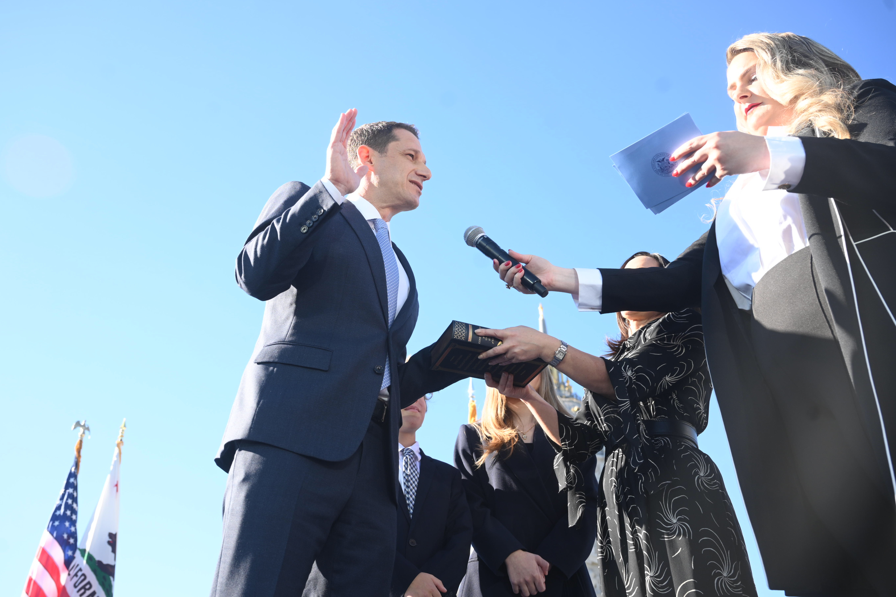 A man in a suit is being sworn in, raising his right hand. A woman holds a microphone to him, and another holds a book. U.S. and California flags are visible.