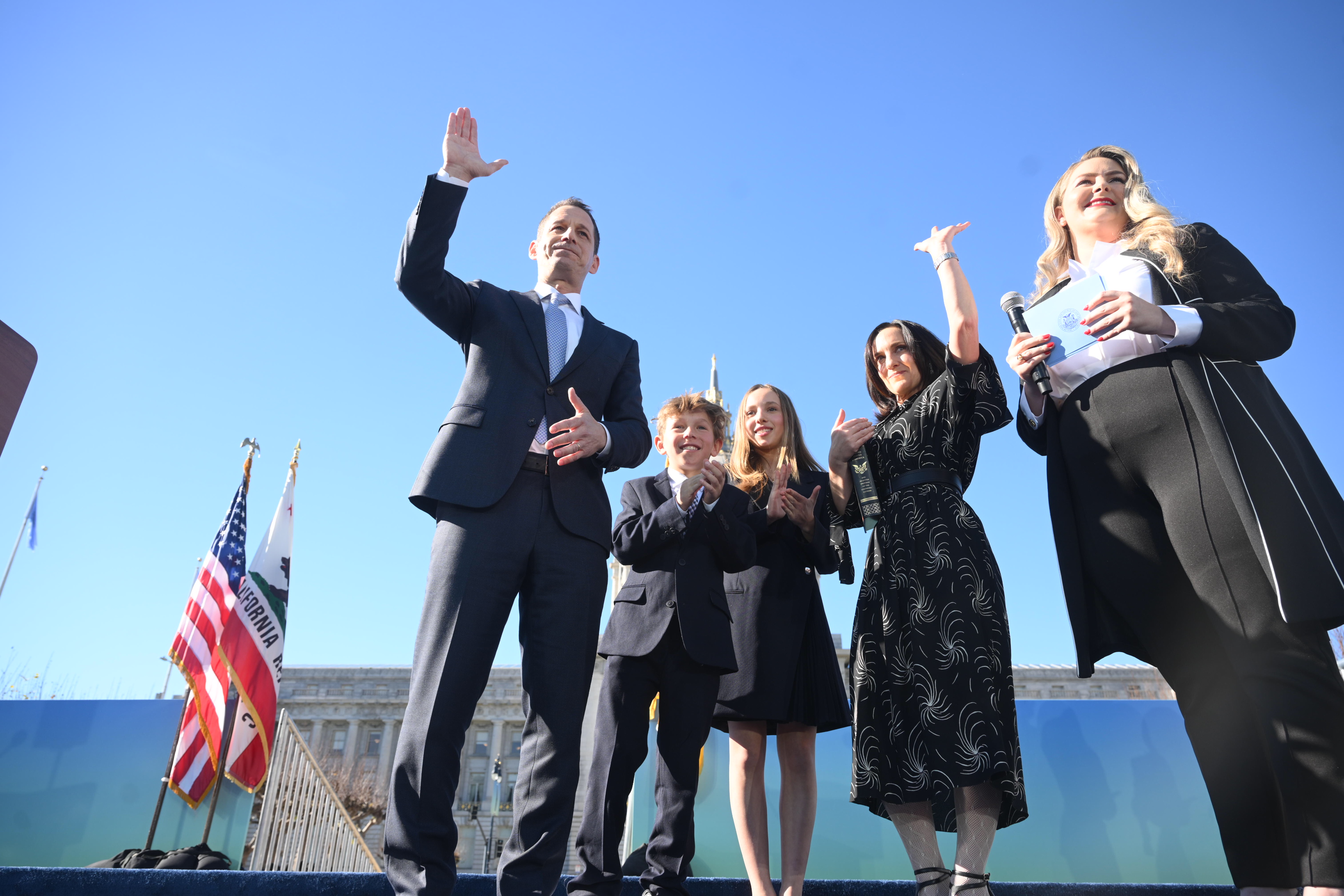 A group of five people, including a child, stand on a stage outdoors. They are dressed formally, waving and smiling. U.S. and California flags are in the background.