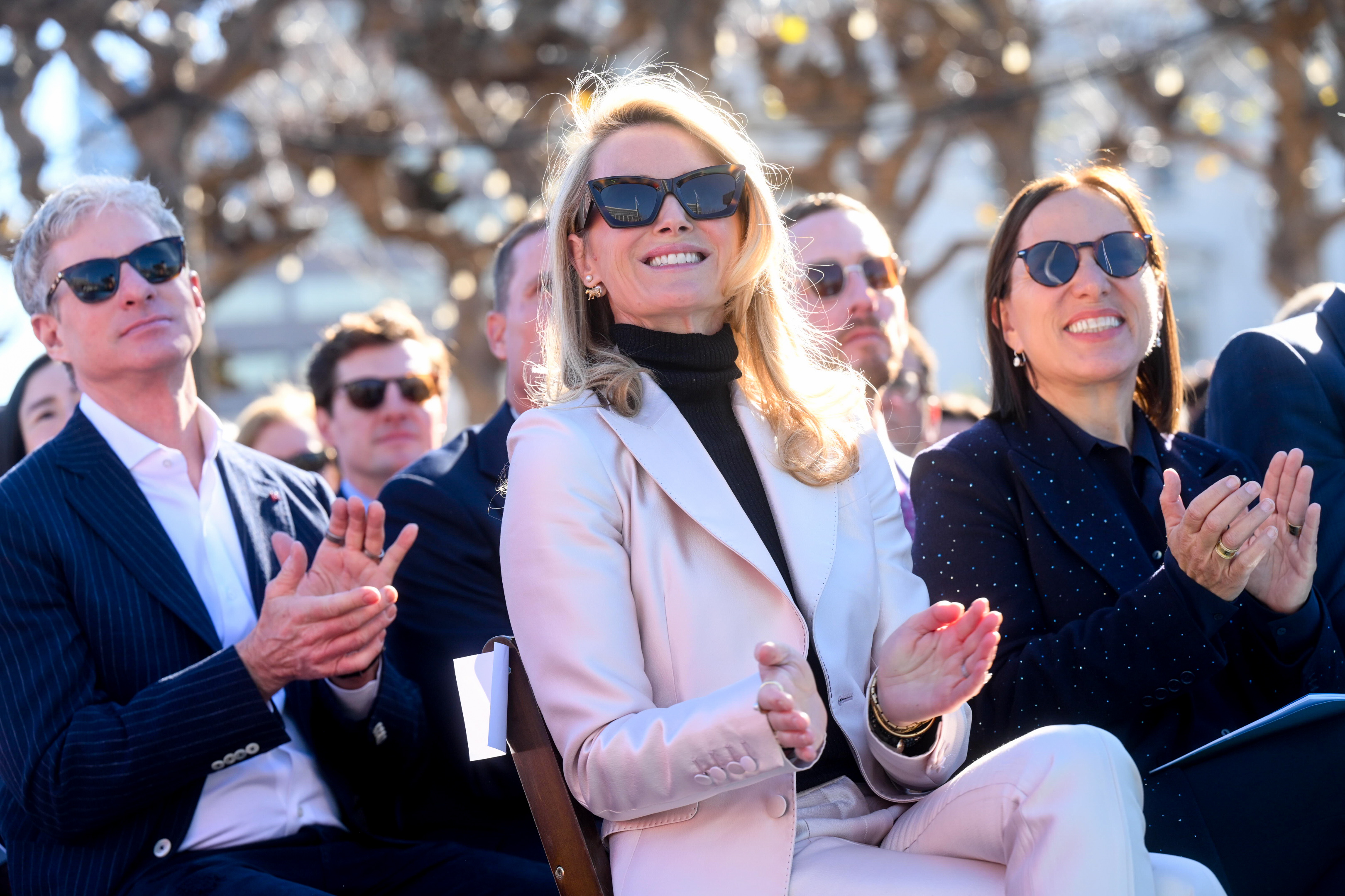 A group of people wearing sunglasses are seated outdoors, smiling and applauding, with trees and sunlight in the background. They appear to be enjoying an event.