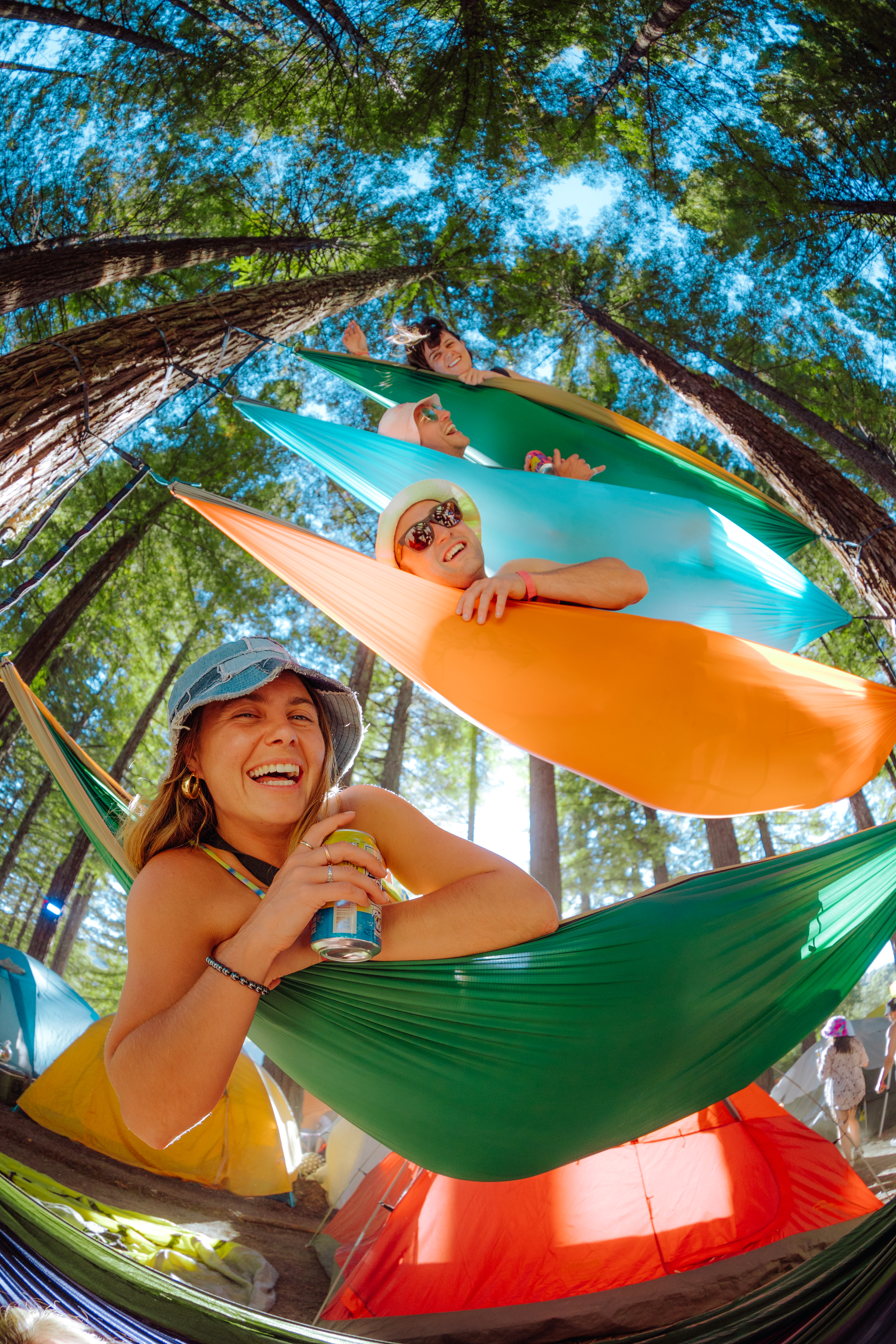 Four people are smiling and relaxing in colorful stacked hammocks under tall trees, with tents visible in the background.