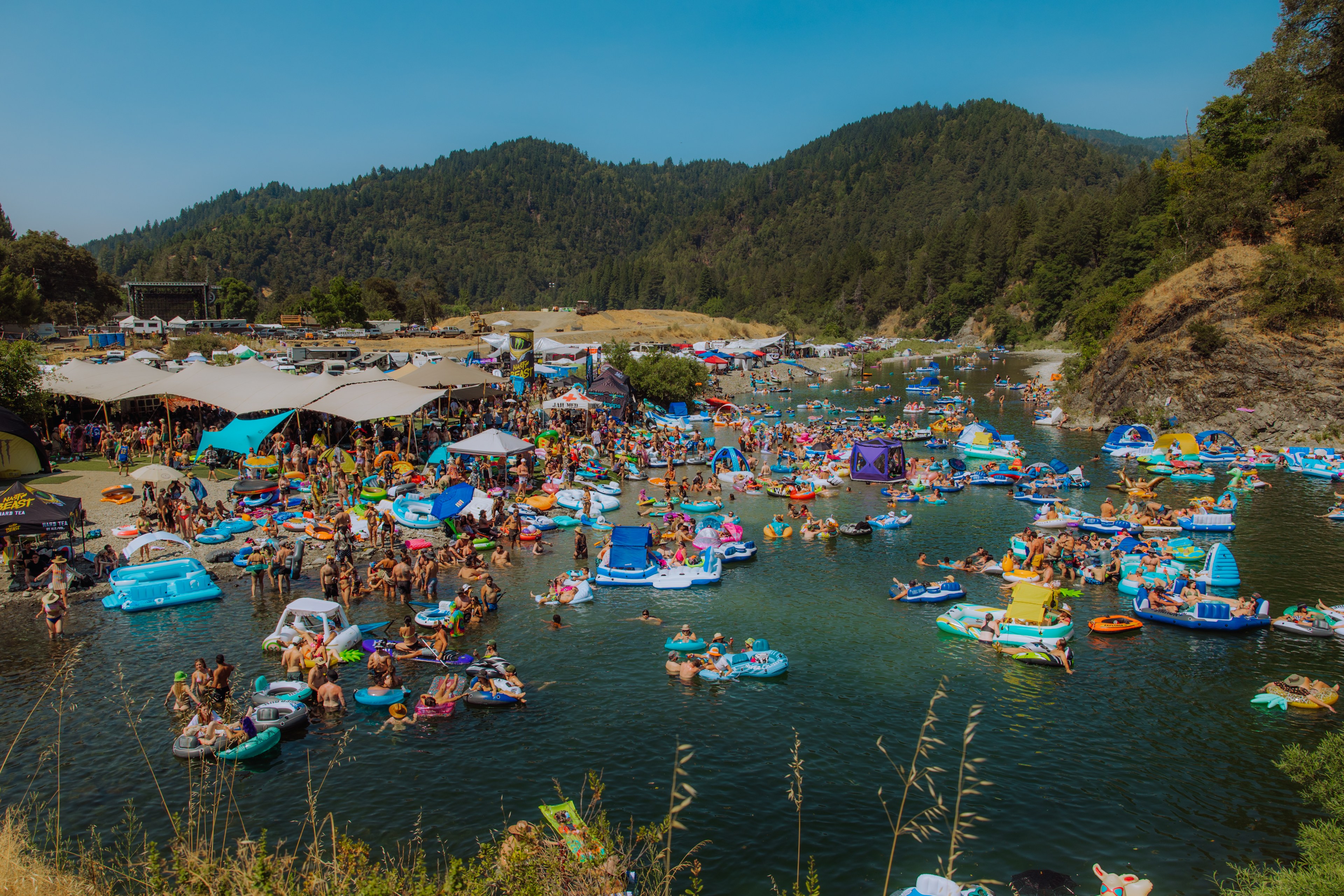 People are relaxing on colorful inflatable rafts in a river, surrounded by a forested landscape. Tents and umbrellas line the shore in the background.