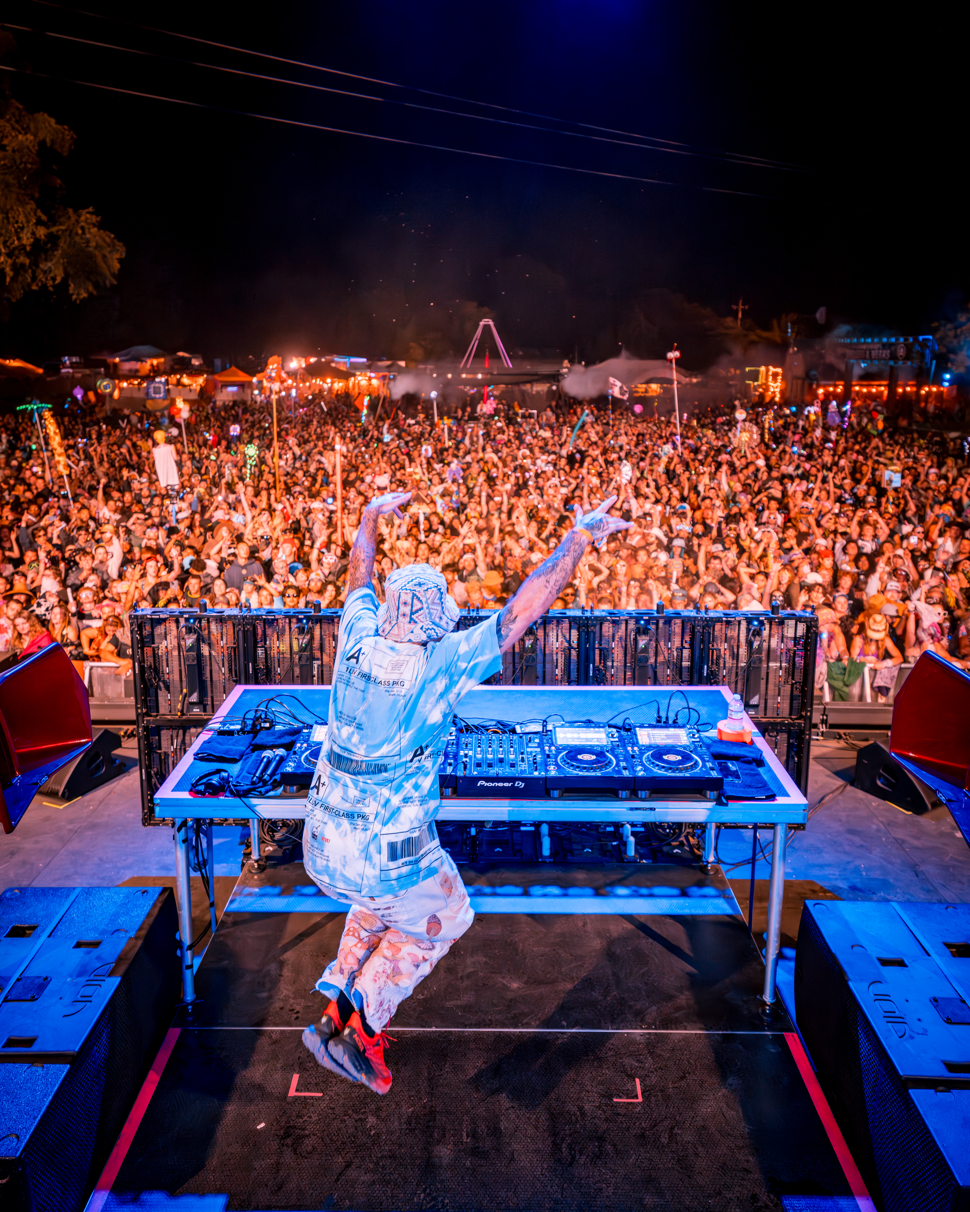 A DJ energetically leaps on stage, facing a vast, vibrant crowd at a night festival. The audience is animated, lights glowing in the background.