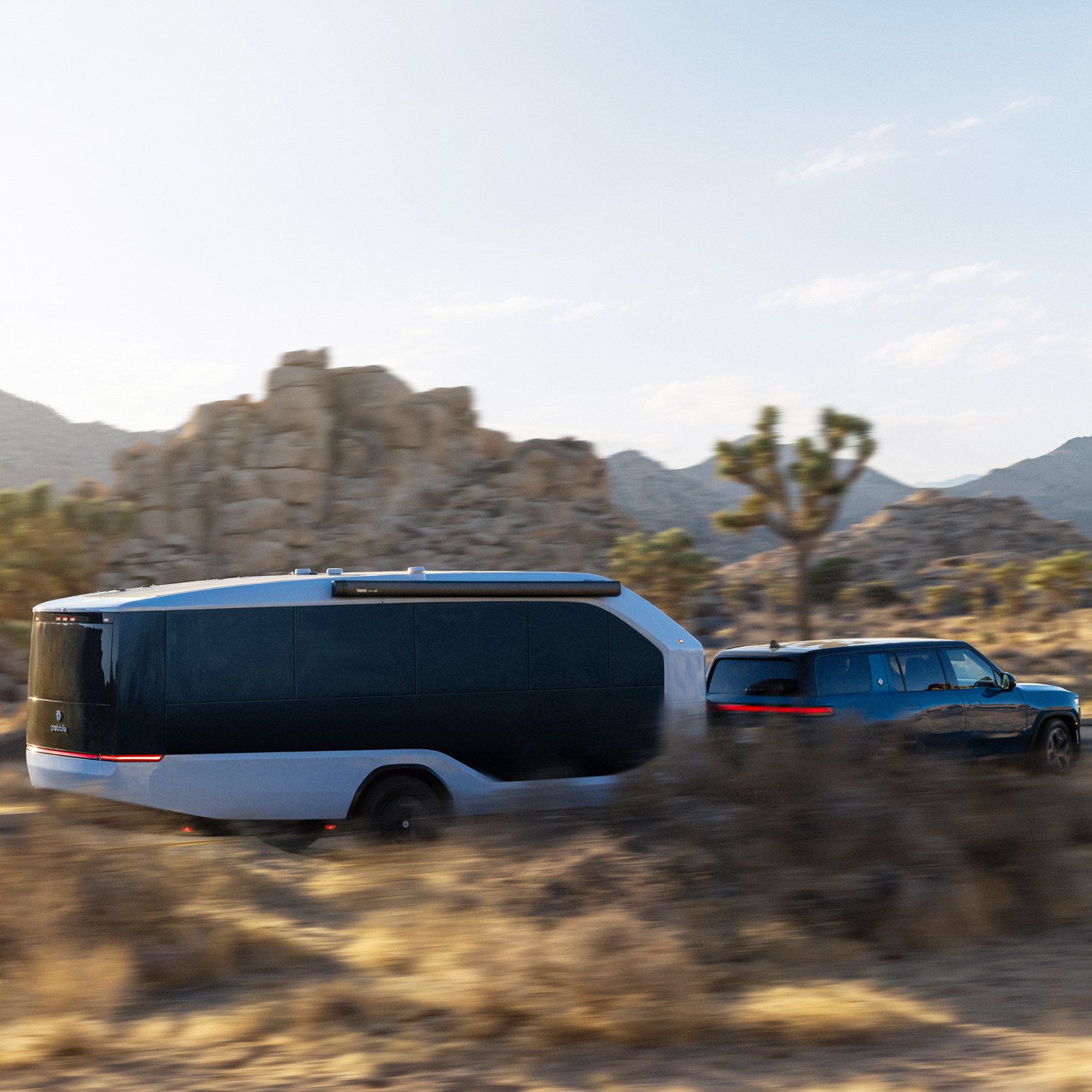 A blue SUV is towing a sleek, modern trailer through a desert landscape with rocky hills and sparse vegetation under a clear sky.