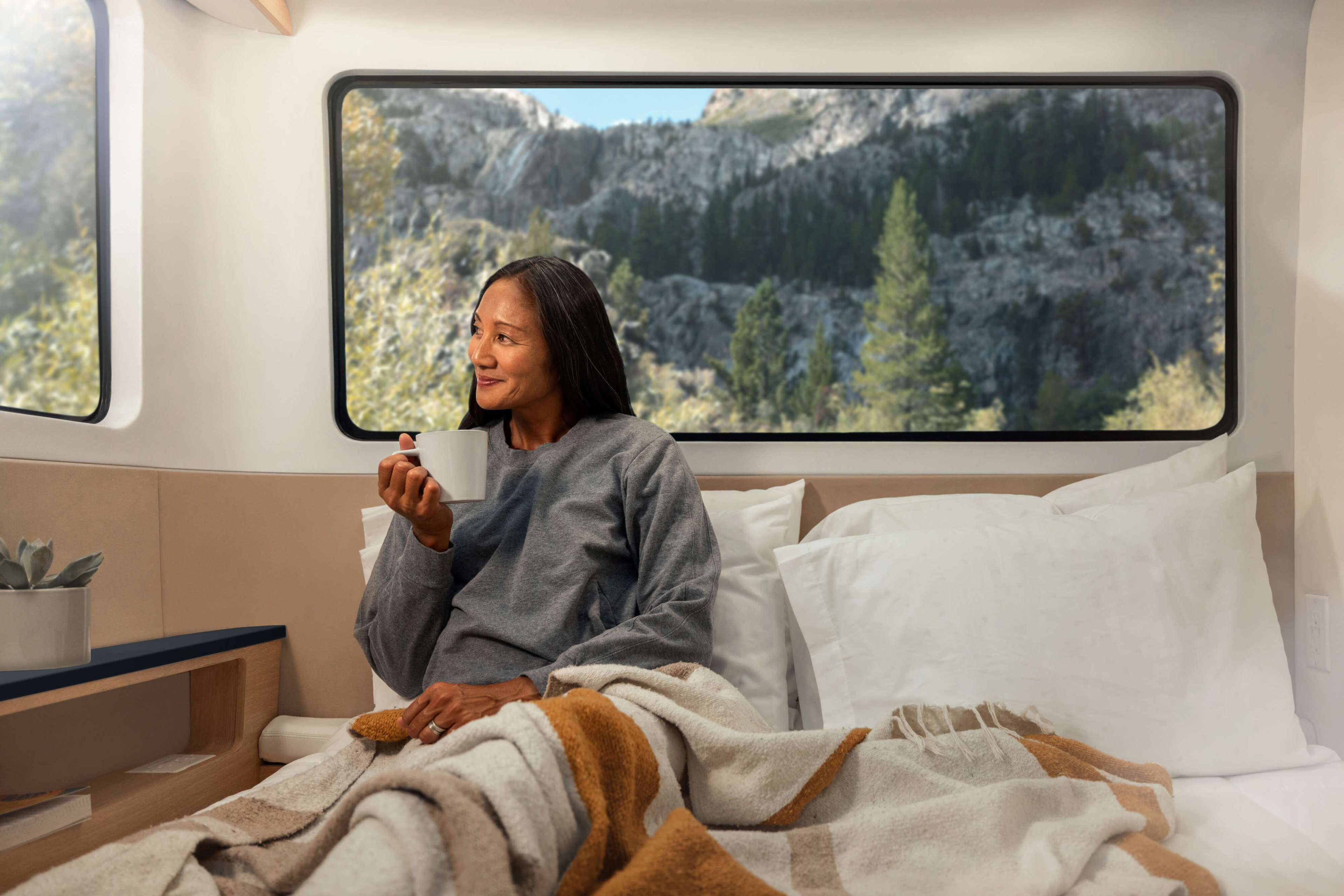 A woman sits on a bed, wrapped in a blanket, sipping from a mug, and gazing out a window at a scenic mountain landscape.