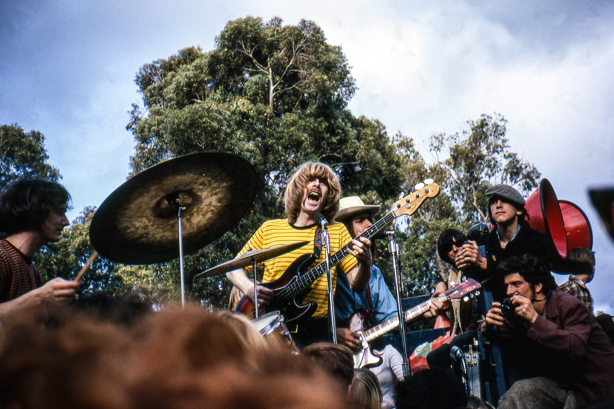 A band performs energetically outdoors, with a drummer, guitarist, and bassist in focus. Trees and a cloudy sky are in the background, and a crowd watches closely.