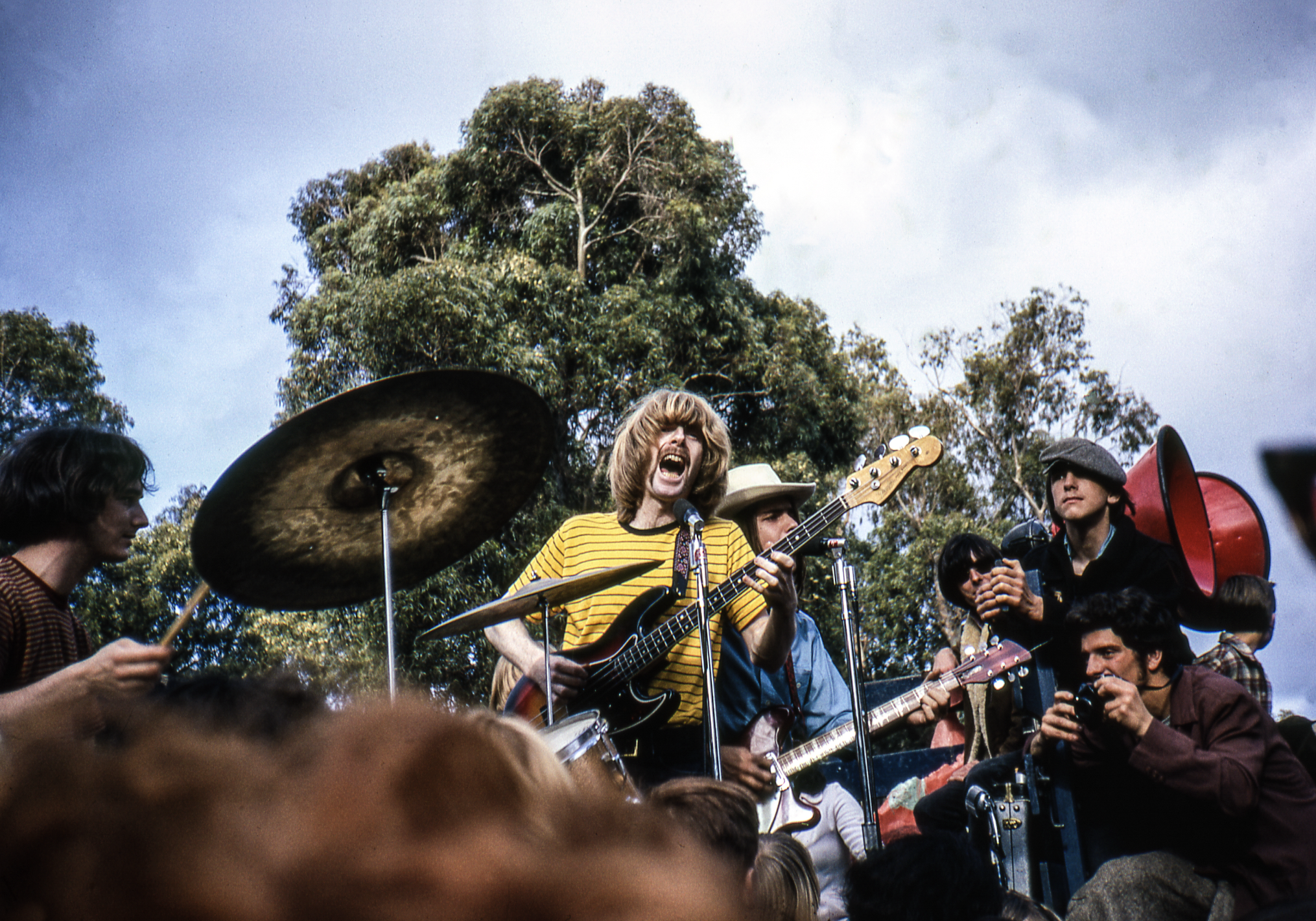 A band performs energetically outdoors, with a drummer, guitarist, and bassist in focus. Trees and a cloudy sky are in the background, and a crowd watches closely.