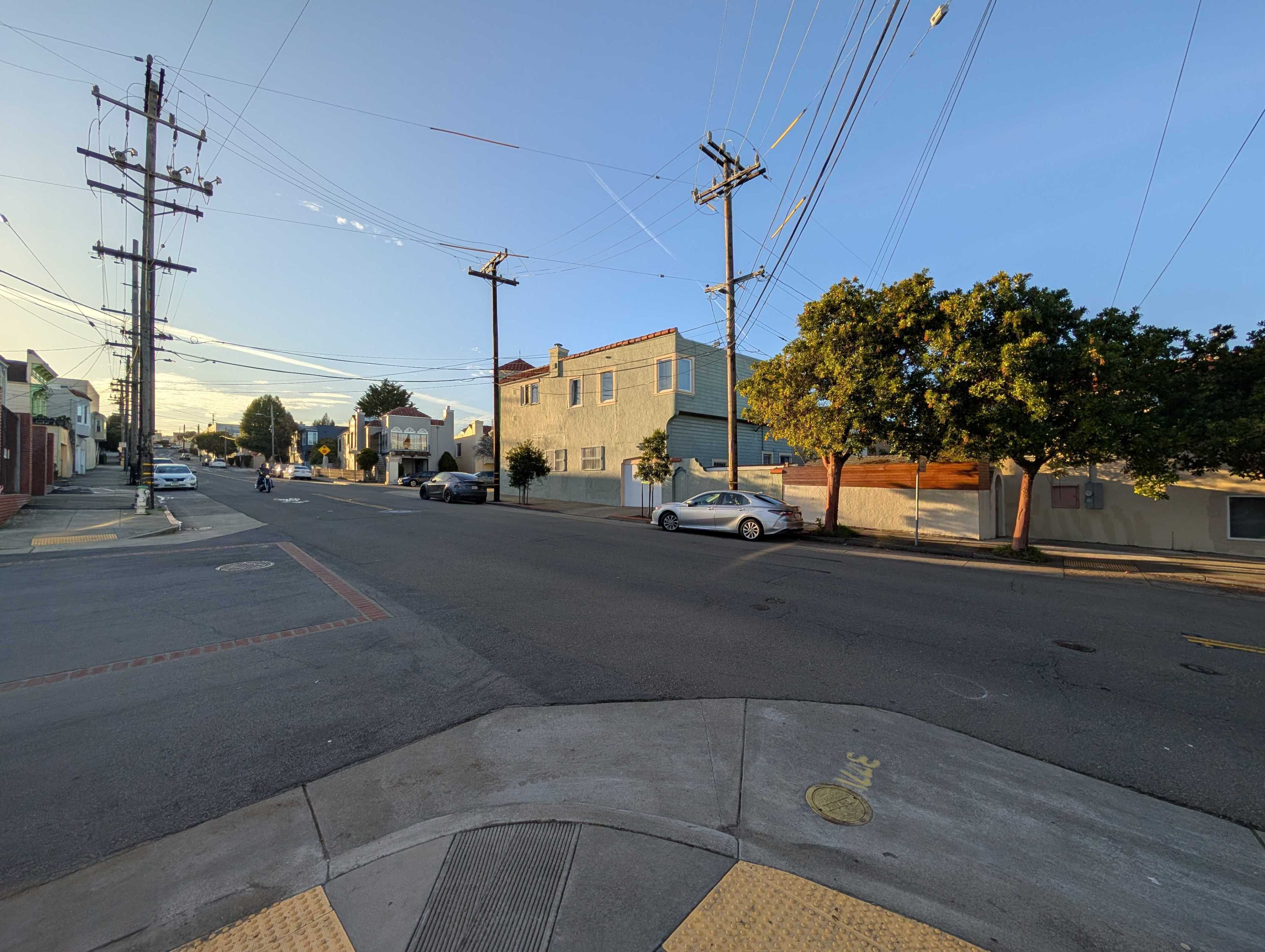 The image shows a quiet urban intersection at sunset, with parked cars, power lines, trees, and residential buildings. The street is mostly empty and peaceful.