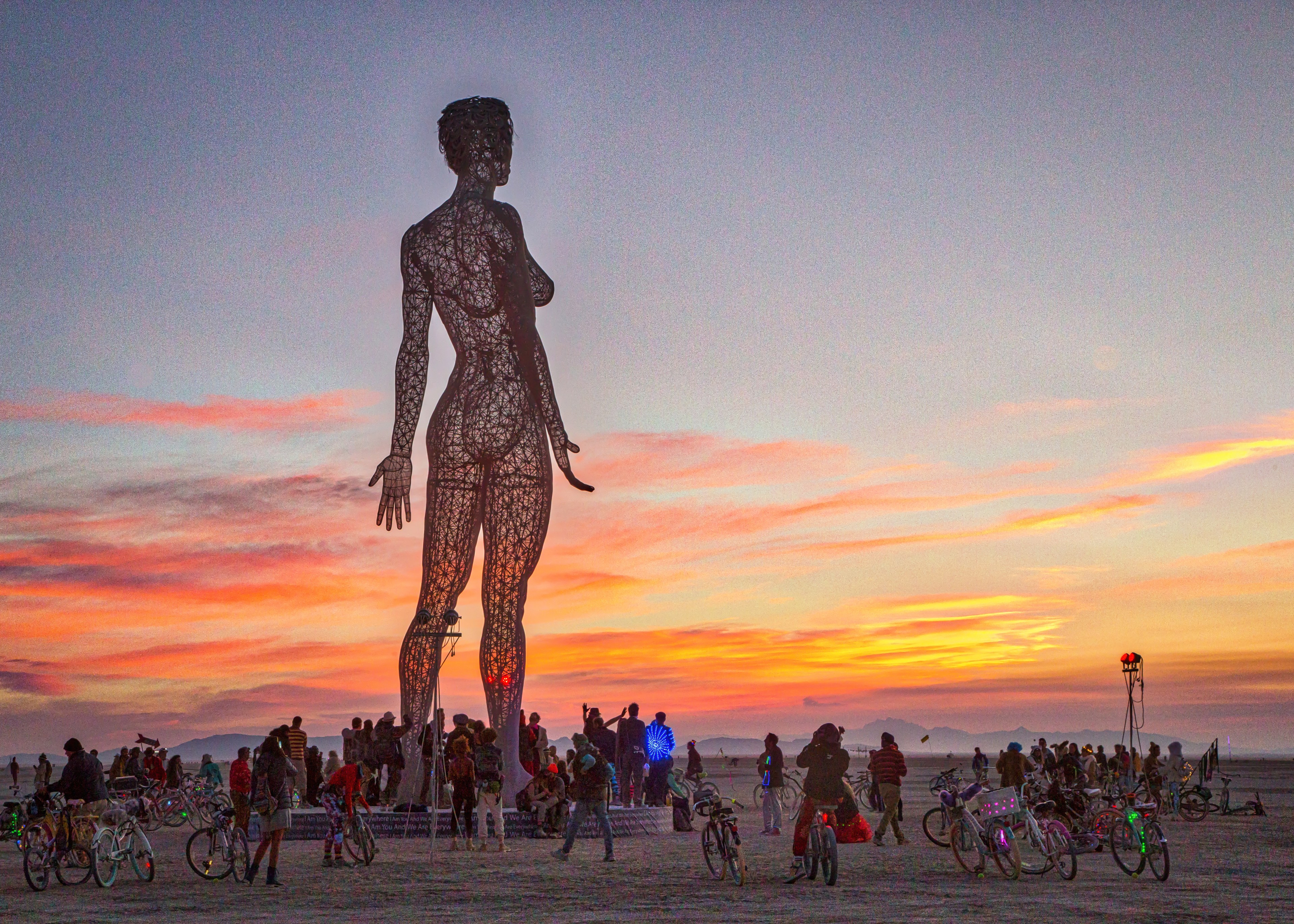 A large, silhouetted sculpture of a woman stands against a vibrant sunset sky. People and bikes surround the sculpture on a flat, open landscape.