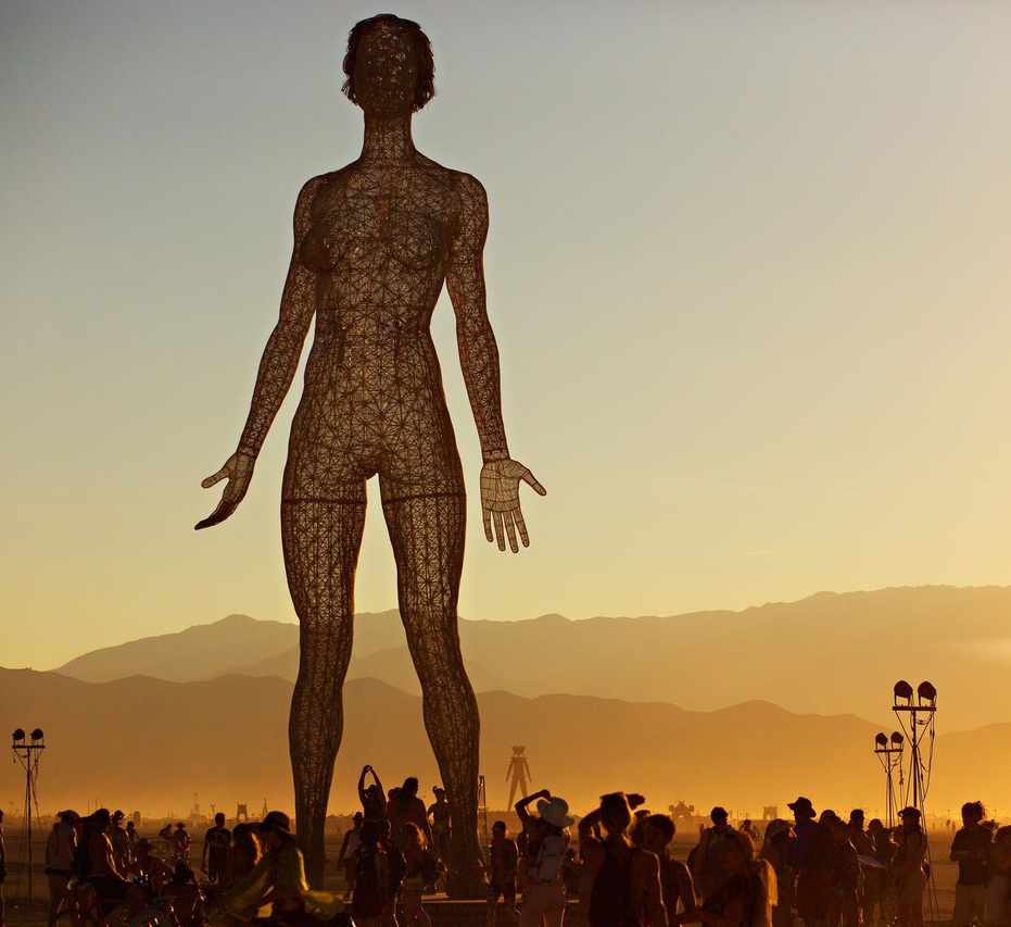 A towering, intricate wire sculpture of a human figure stands against a dusky sky, surrounded by a crowd of people and distant mountains.