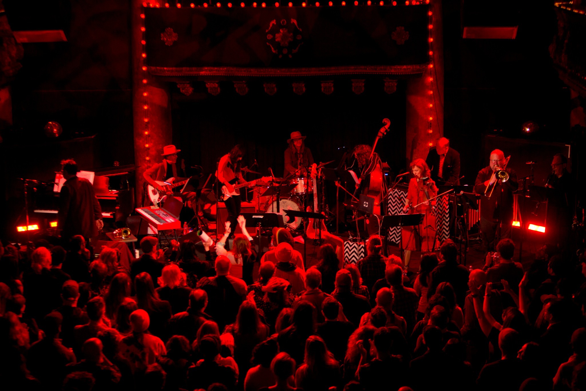 A lively band performs on stage under red lighting, with a diverse range of instruments surrounded by a large, engaged audience.