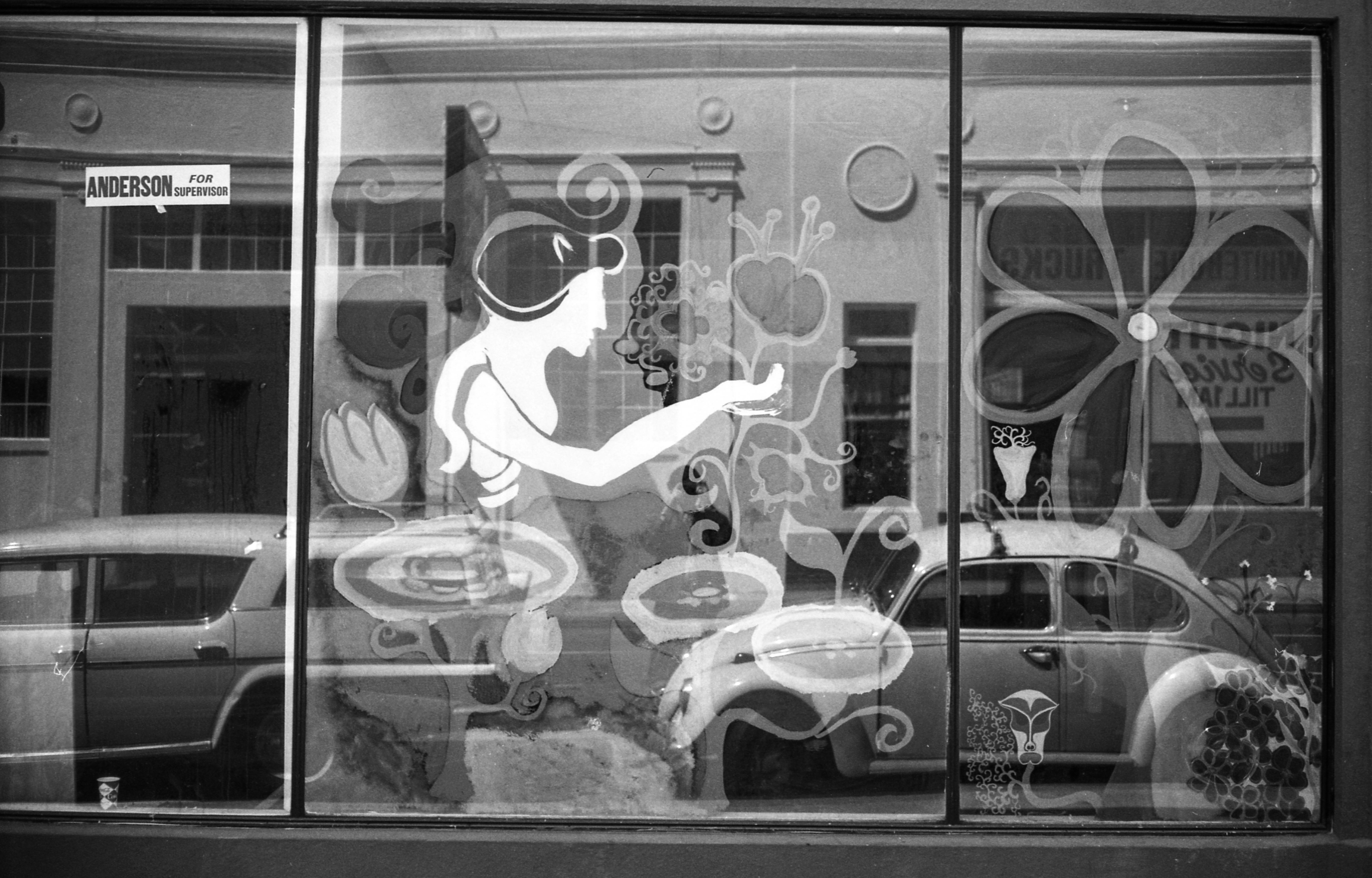 The image shows a storefront window with an artistic depiction of a figure surrounded by flowers and swirls. Reflecting on the glass are vintage cars and a street scene.