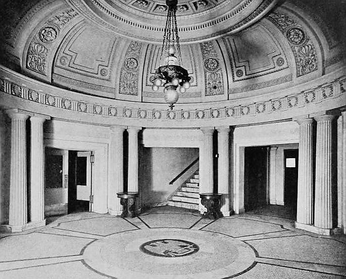 The image shows a circular room with a domed ceiling adorned with intricate designs, flanked by columns. A grand chandelier hangs at the center above patterned flooring.