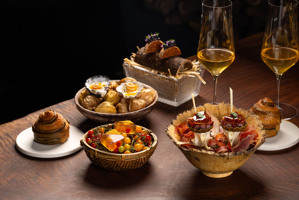 The image shows an elegant spread of gourmet dishes, including pastries, intricate appetizers, and colorful garnishes, accompanied by two glasses of white wine.